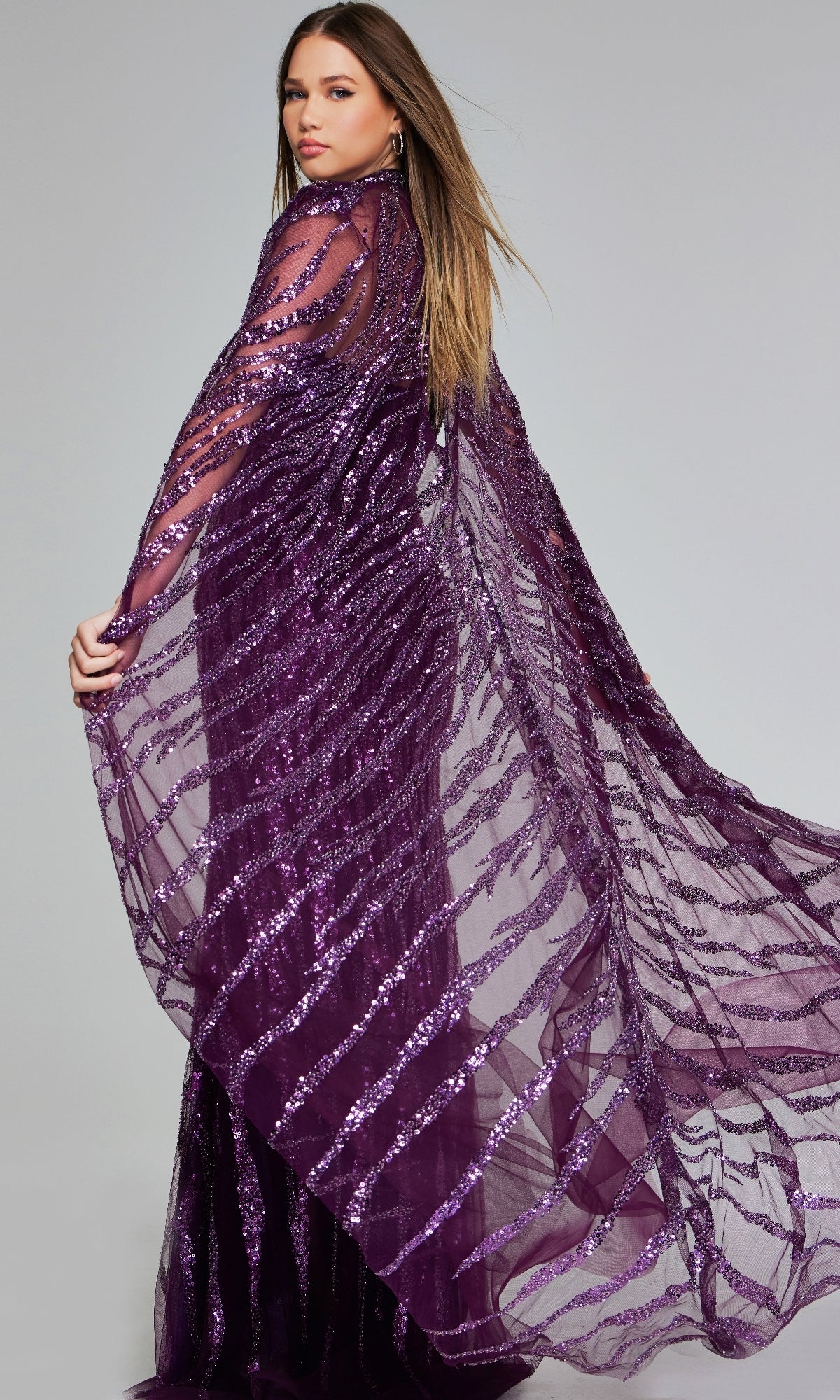 Woman poses in the Jovani 39046 purple sequin pageant dress—long, sheer, with stripe details and flowing cape sleeve—creating an elegant special occasion look against a plain backdrop.