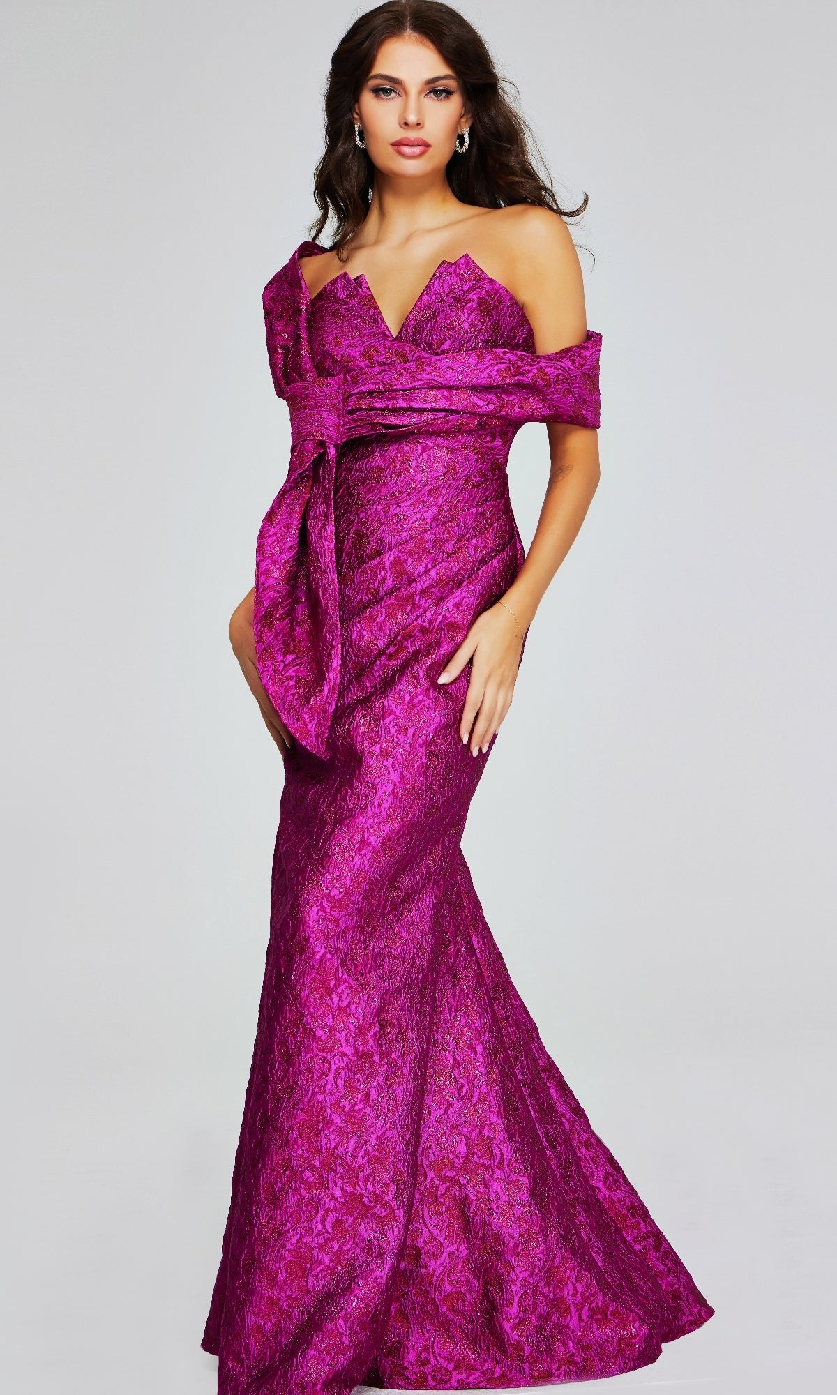 A woman poses elegantly against a plain light background in the Jovani 39114, a long formal off-shoulder magenta gown featuring a textured floral pattern and mermaid silhouette.