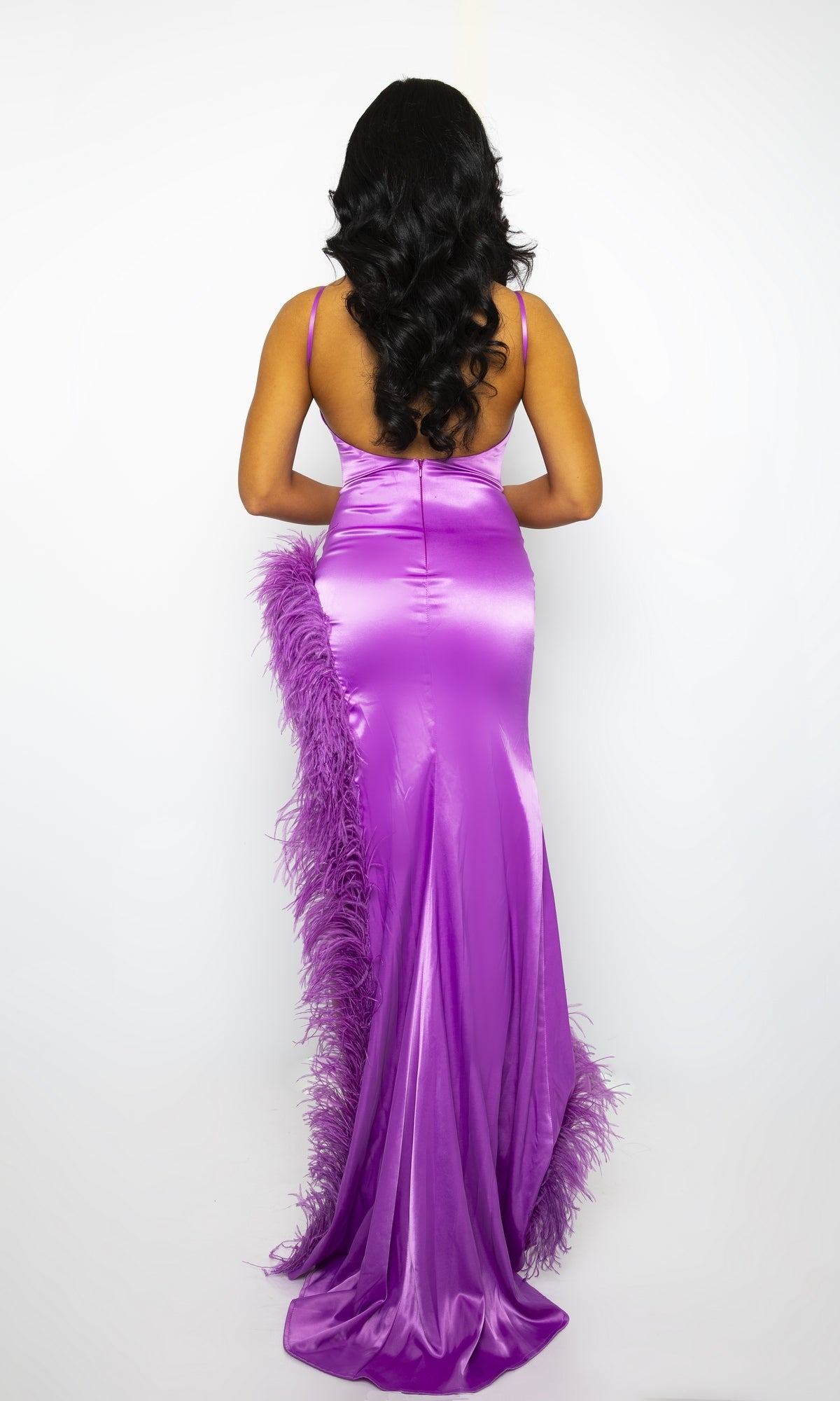 A person with long, dark, wavy hair models the Ava Presley Long Prom Dress 39224—a shiny purple open-back formal gown with feather trim—viewed from the back against a plain white background.