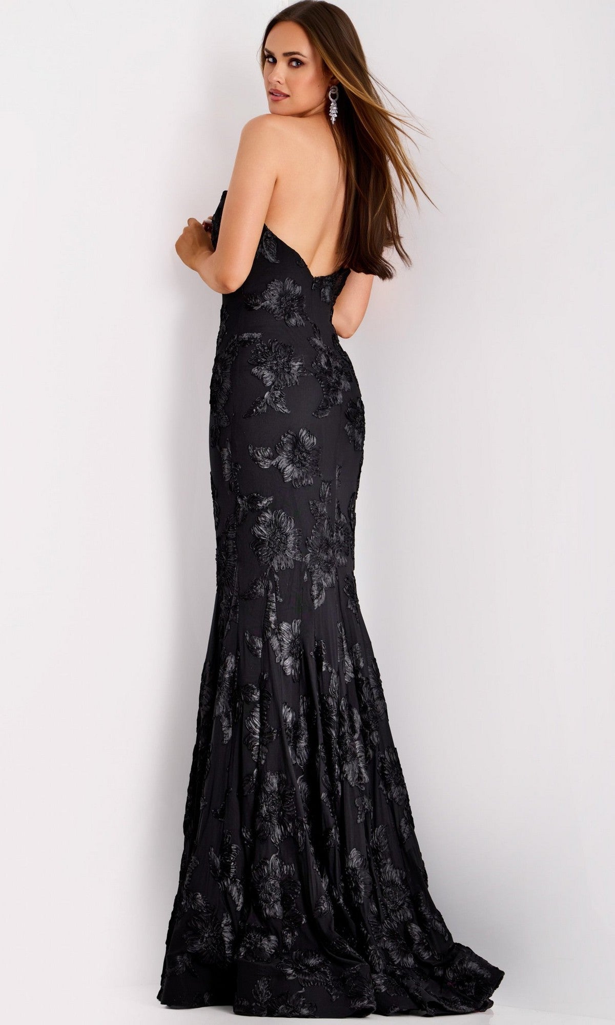 A woman models the Jovani 39267 long formal dress—a strapless, black, floor-length designer evening gown with a low back, elegant floral-patterned design, and a subtle mermaid silhouette—as she stands looking over her shoulder against a plain background.