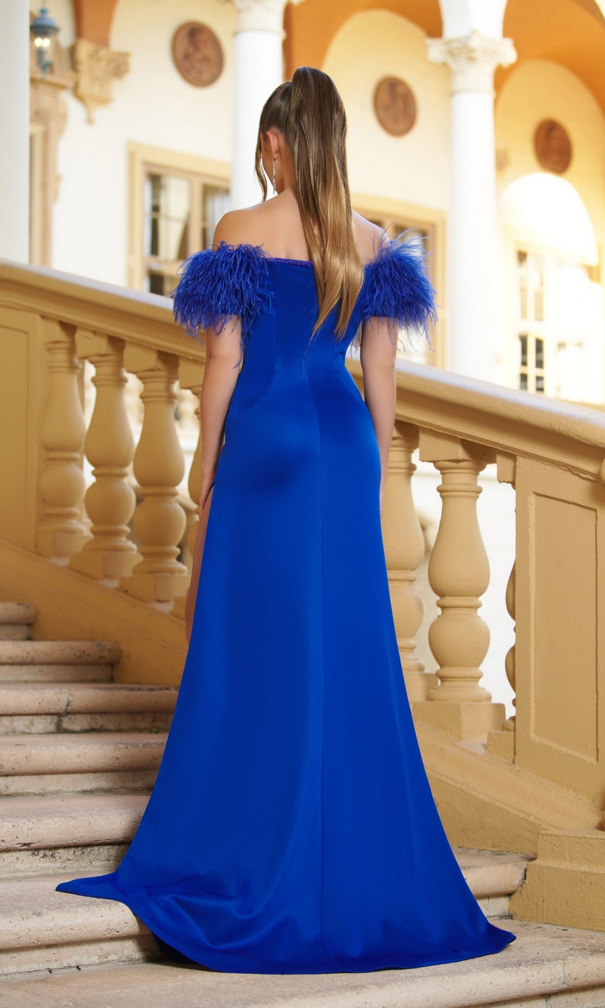 A woman in the Ava Presley 39268 Feathered Long Prom Dress, featuring royal blue off-the-shoulder feathered banded sleeves, stands on a staircase with her back to the camera.