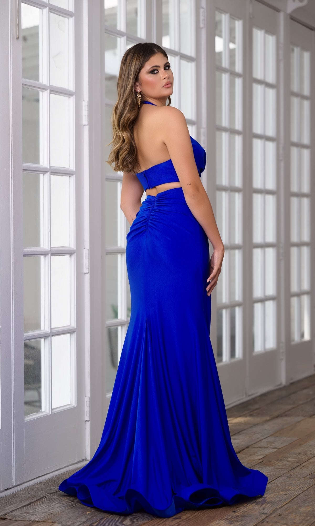 A woman models the Ava Presley 39271 Cut-Out Long Halter Prom Dress, a fitted, floor-length royal blue gown. She poses gracefully indoors, facing away with a slight turn toward the camera in front of white-paneled glass doors.