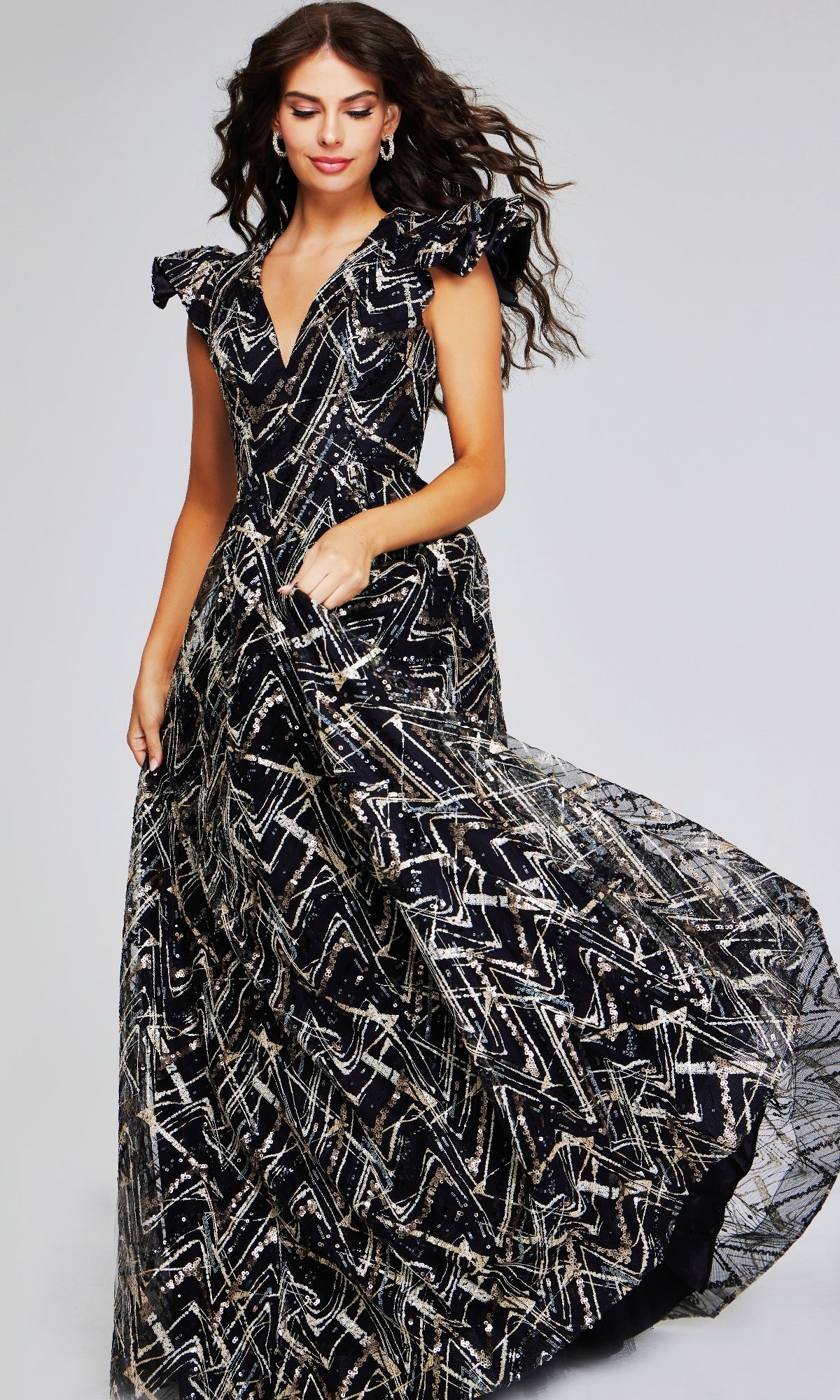 Woman in the Jovani 39274 long formal dress, featuring black and gold metallic geometric patterns and ruffled cap sleeves, stands against a plain background.