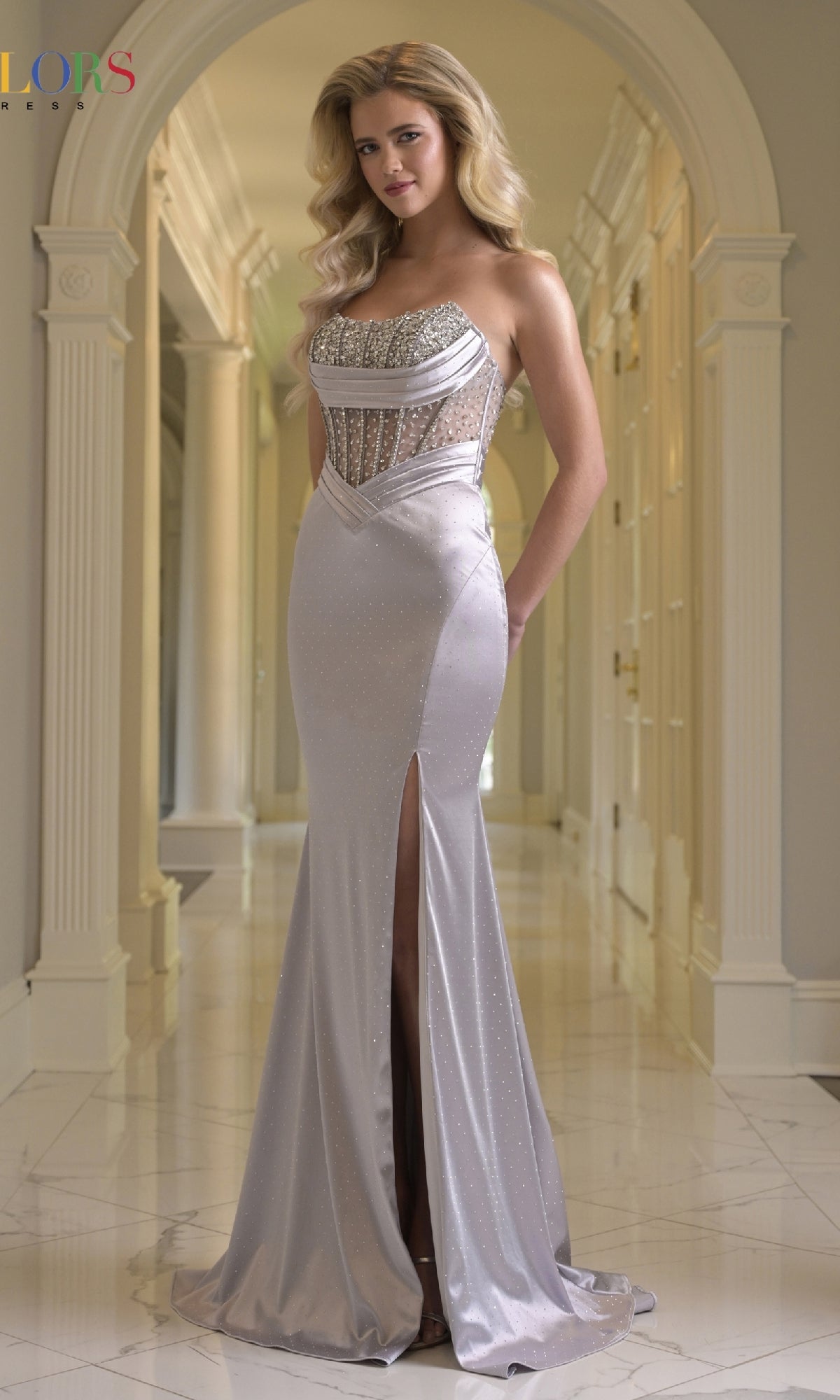A woman stands in a hallway wearing the Beaded-Corset Satin Prom Gown: Colors Dress 3932 in silver, a fitted, floor-length gown featuring a thigh-high slit and stunning beaded corset detailing.