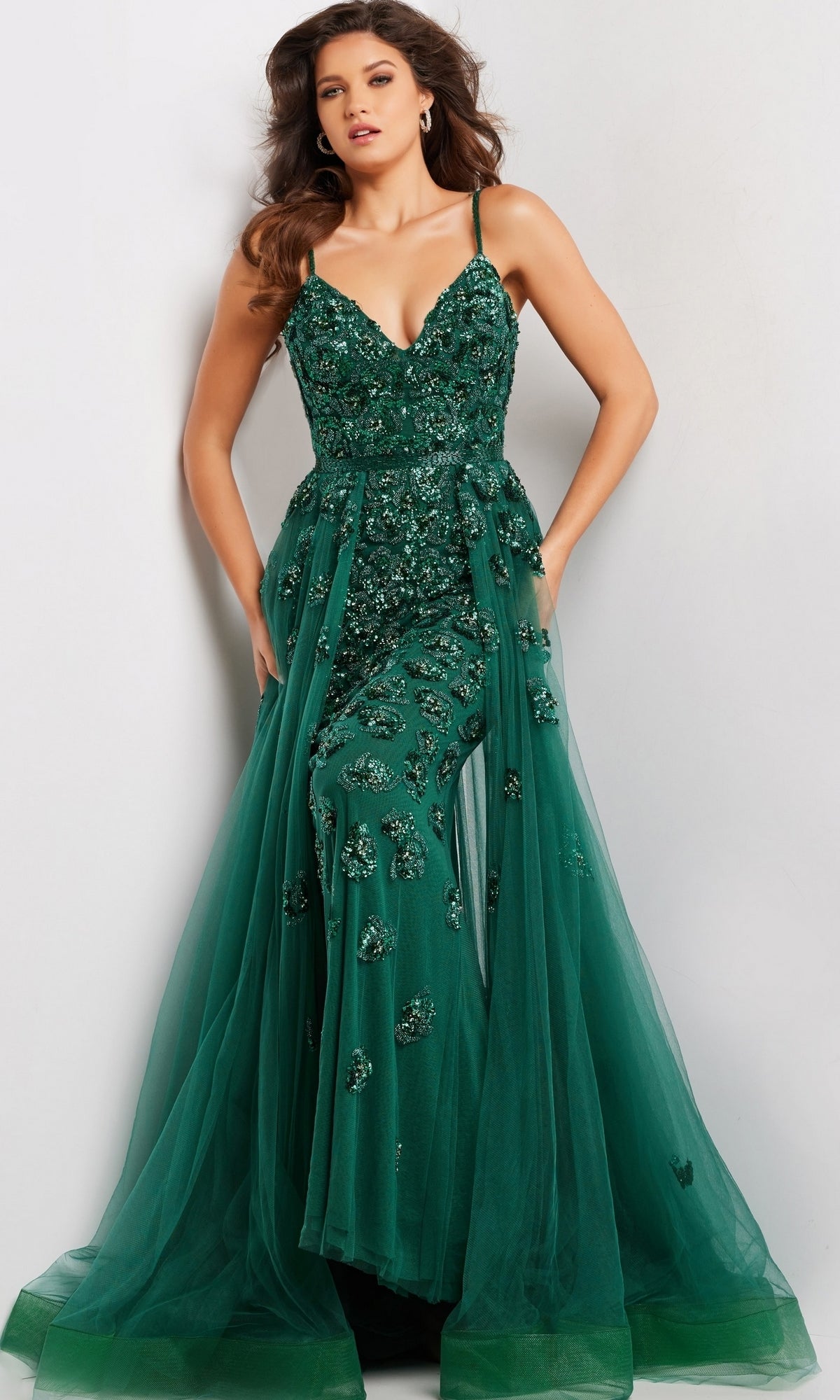 A woman models the Long Prom Dress 39434 by Jovani—a deep green formal gown with thin straps, beaded details, floral embellishments, and a flowing tulle A-line overskirt—posed against a plain background.