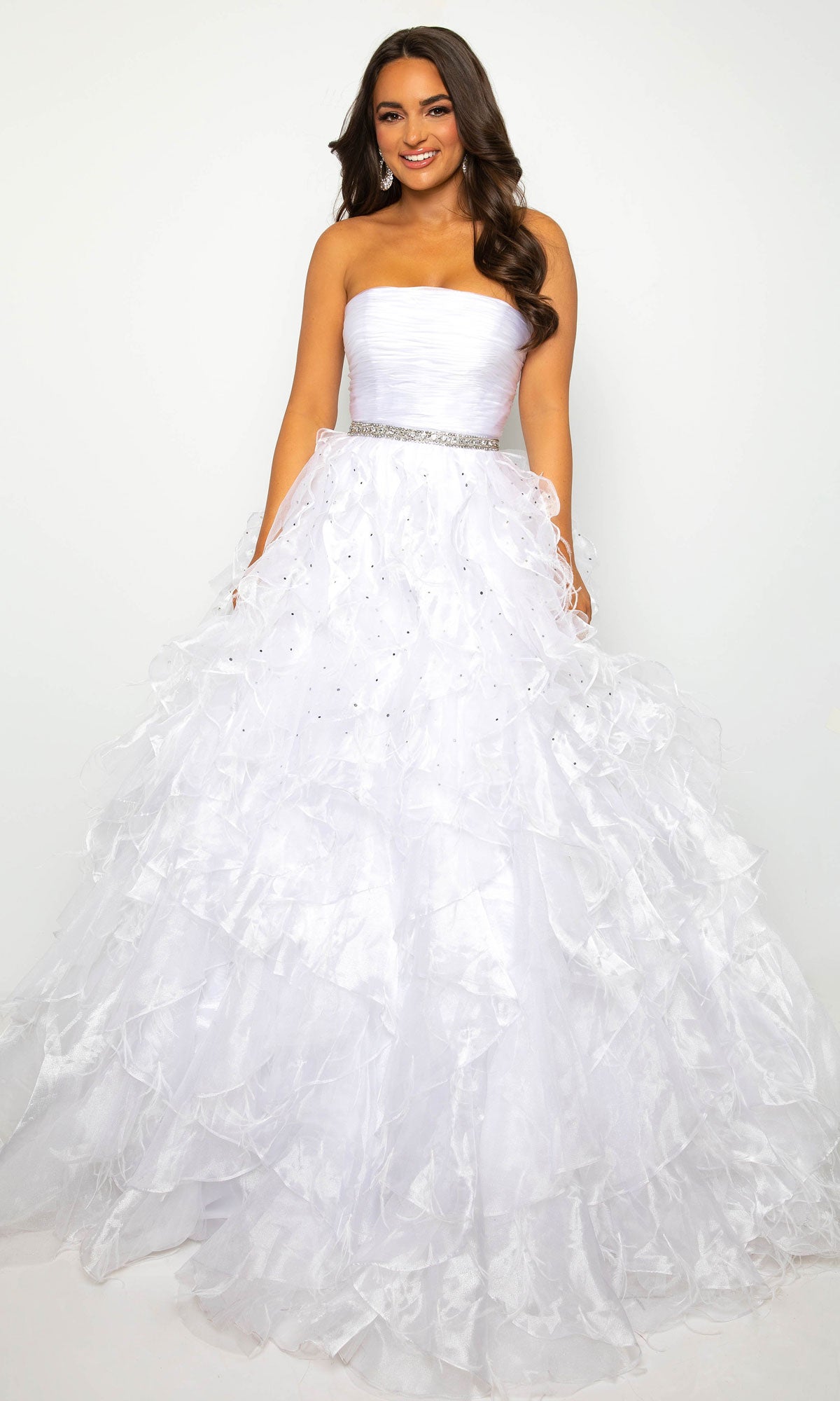 A woman smiles at the camera in the Strapless Ruffle Prom Gown: Ava Presley 39555, featuring a jeweled waistband and ruffled A-line skirt, standing against a plain white background.