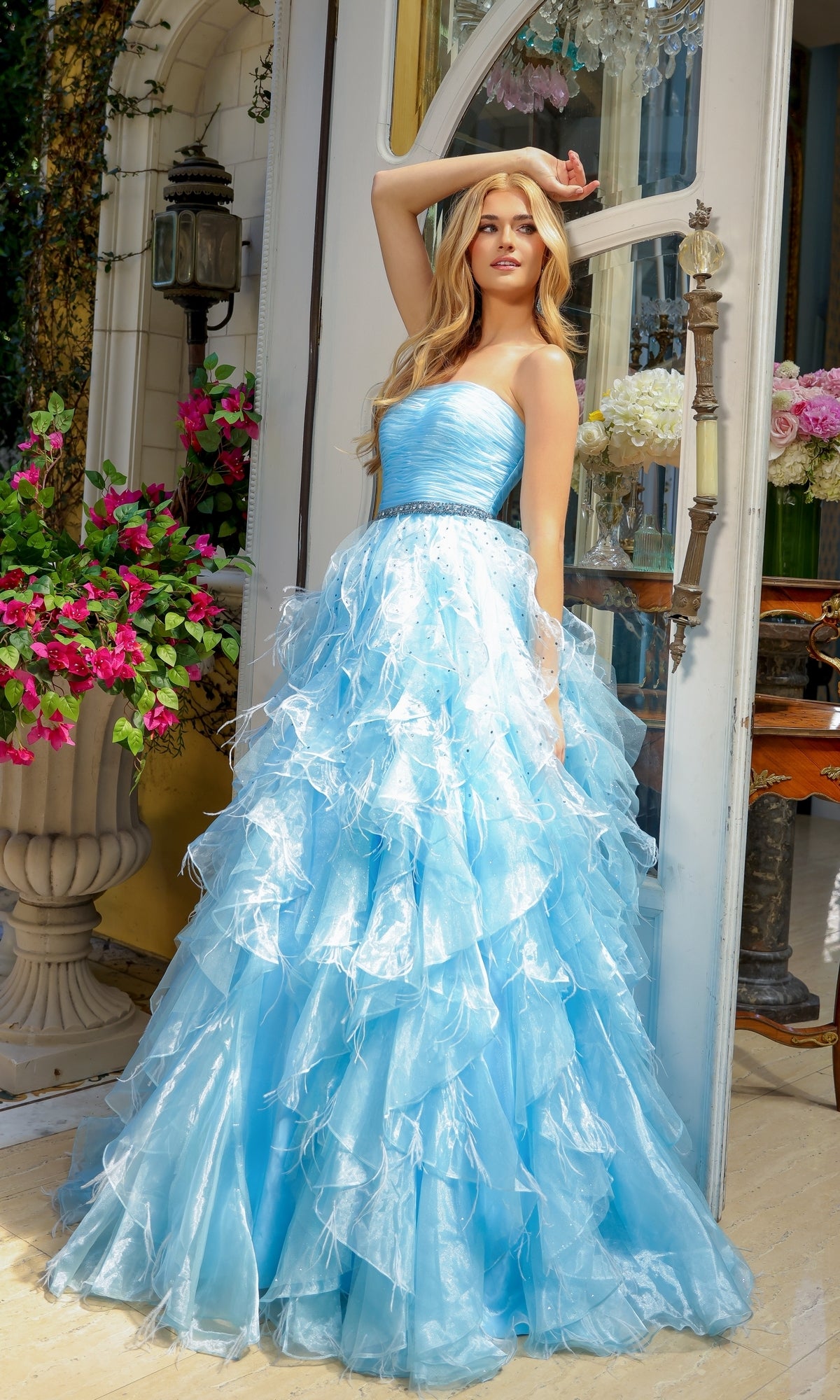 A young woman wearing the Ava Presley 39555 Strapless Ruffle Prom Gown stands by a glass door, surrounded by flowers and outdoor decor, her elegant A-line gown flowing around her.