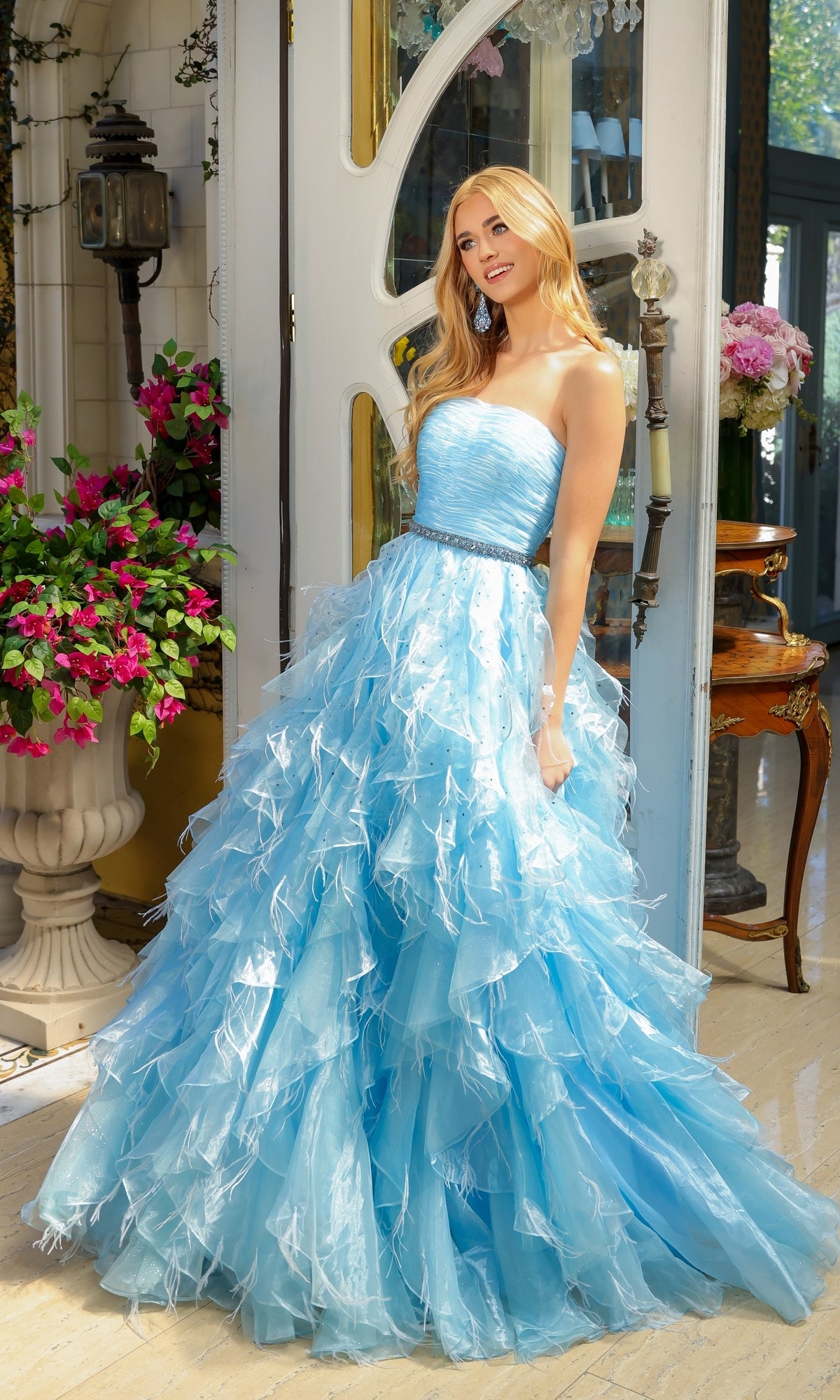 A woman in the Ava Presley 39555 Strapless Ruffle Prom Gown stands indoors by a door, surrounded by pink flowers and ornate furniture, wearing a light blue, ruffled A-line dress.