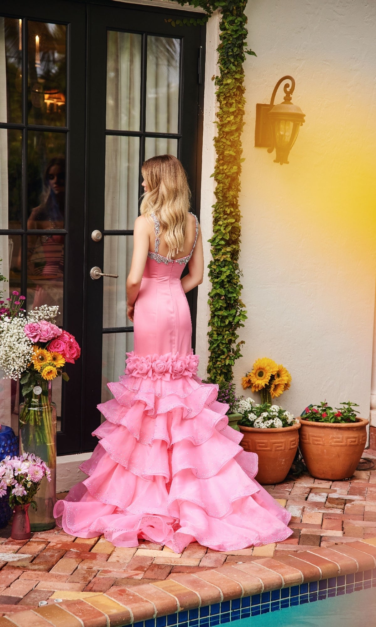 A woman wearing the Ava Presley 39566 Ruffle Mermaid Prom Dress poses by a glass door near potted sunflowers and flowers on a brick patio beside a pool.