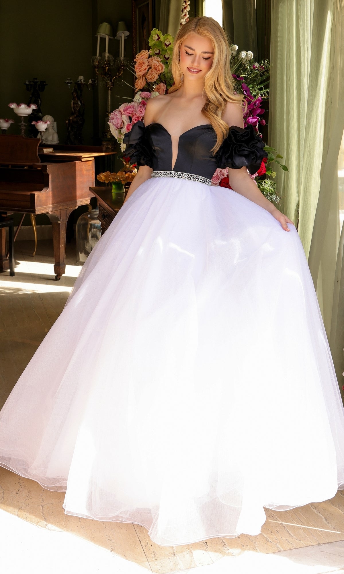 A woman smiles indoors, holding out the sides of her Long Prom Dress 39581 by Ava Presley—an off-the-shoulder puff sleeve top with a full white tulle A-line skirt. Elegant flowers and a piano decorate the background.