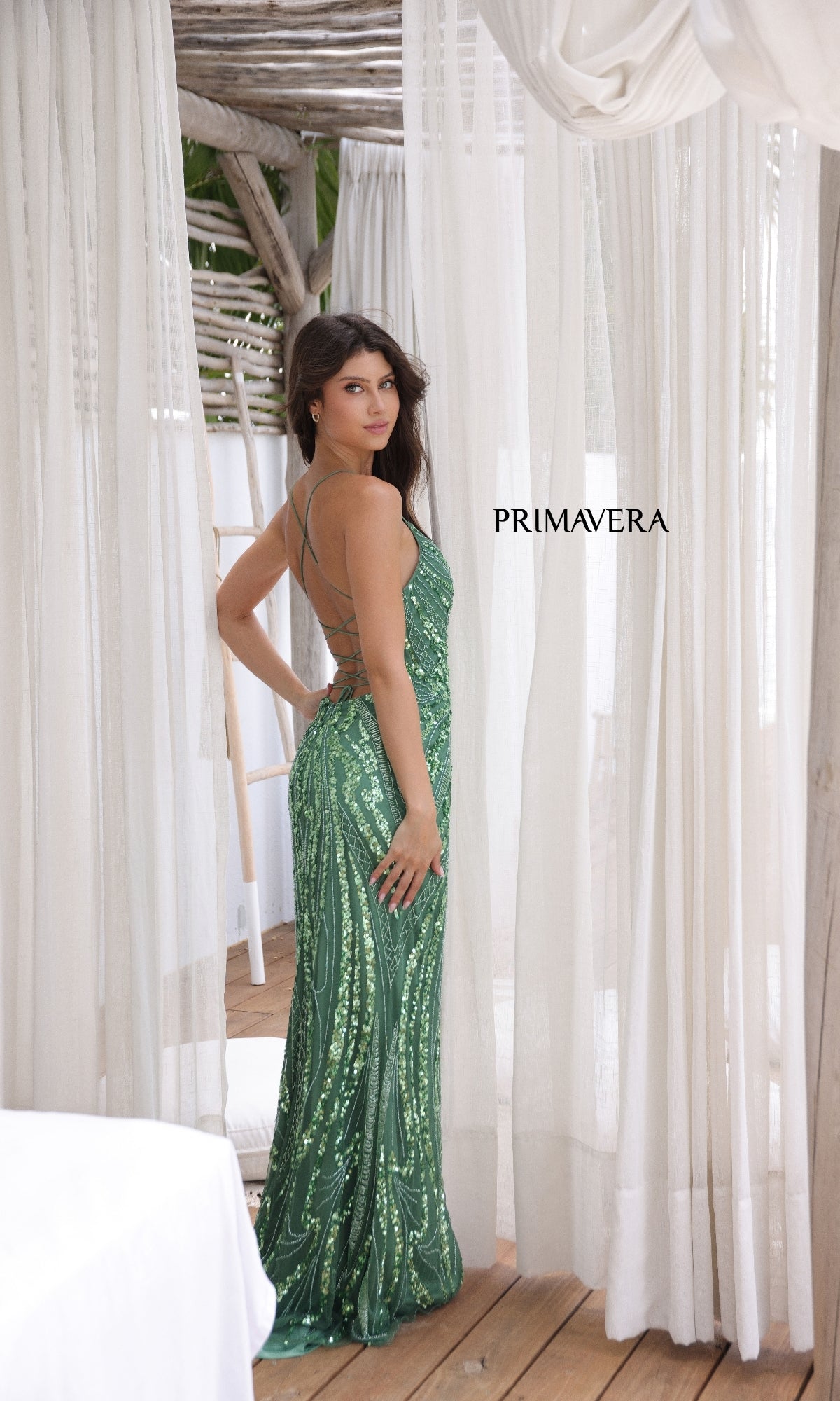 A woman wears the Strappy-Back Long Sequin Prom Dress 3958 in green, standing on a wooden deck with white curtains, glancing over her shoulder.