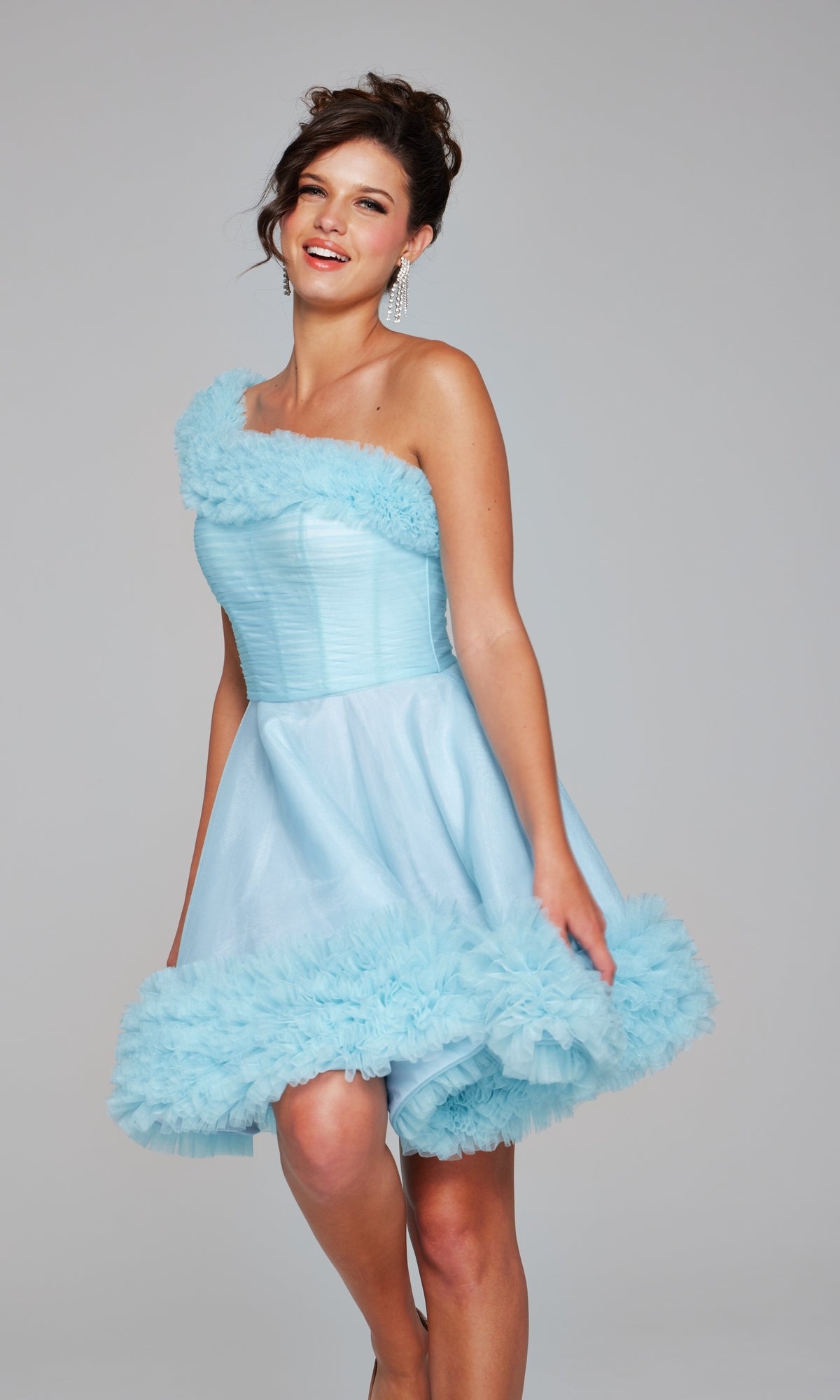 Woman wearing the Jovani 39665 One-Shoulder Short Ruffle Hoco Dress in light blue, featuring ruffled details on the neckline and hem, standing against a plain gray background.