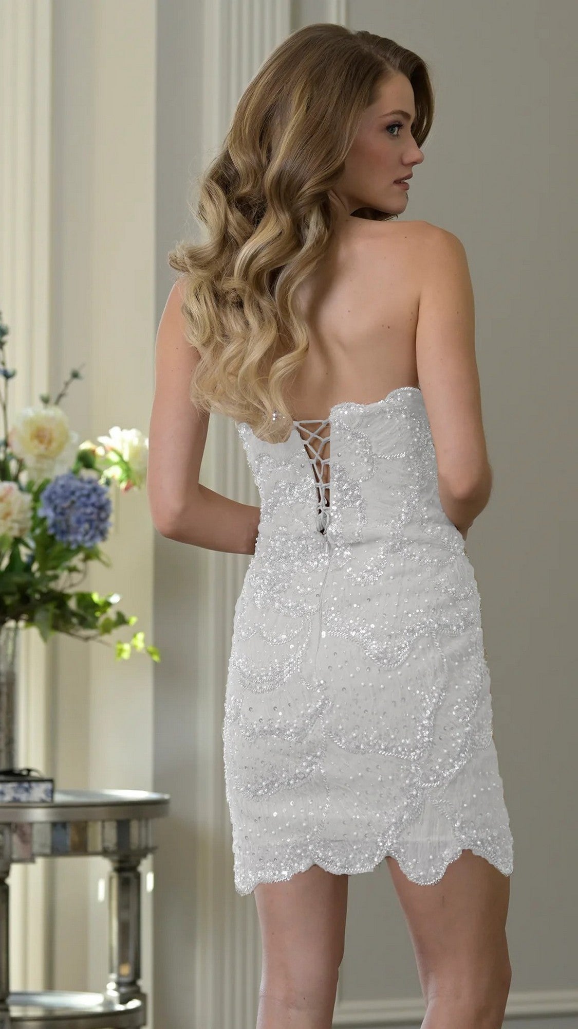 A woman with wavy hair wears the Short Homecoming Dress: Colors Dress 3970—a strapless sequin dress with a lace-up back—standing indoors near a table with flowers, making an elegant choice for homecoming.