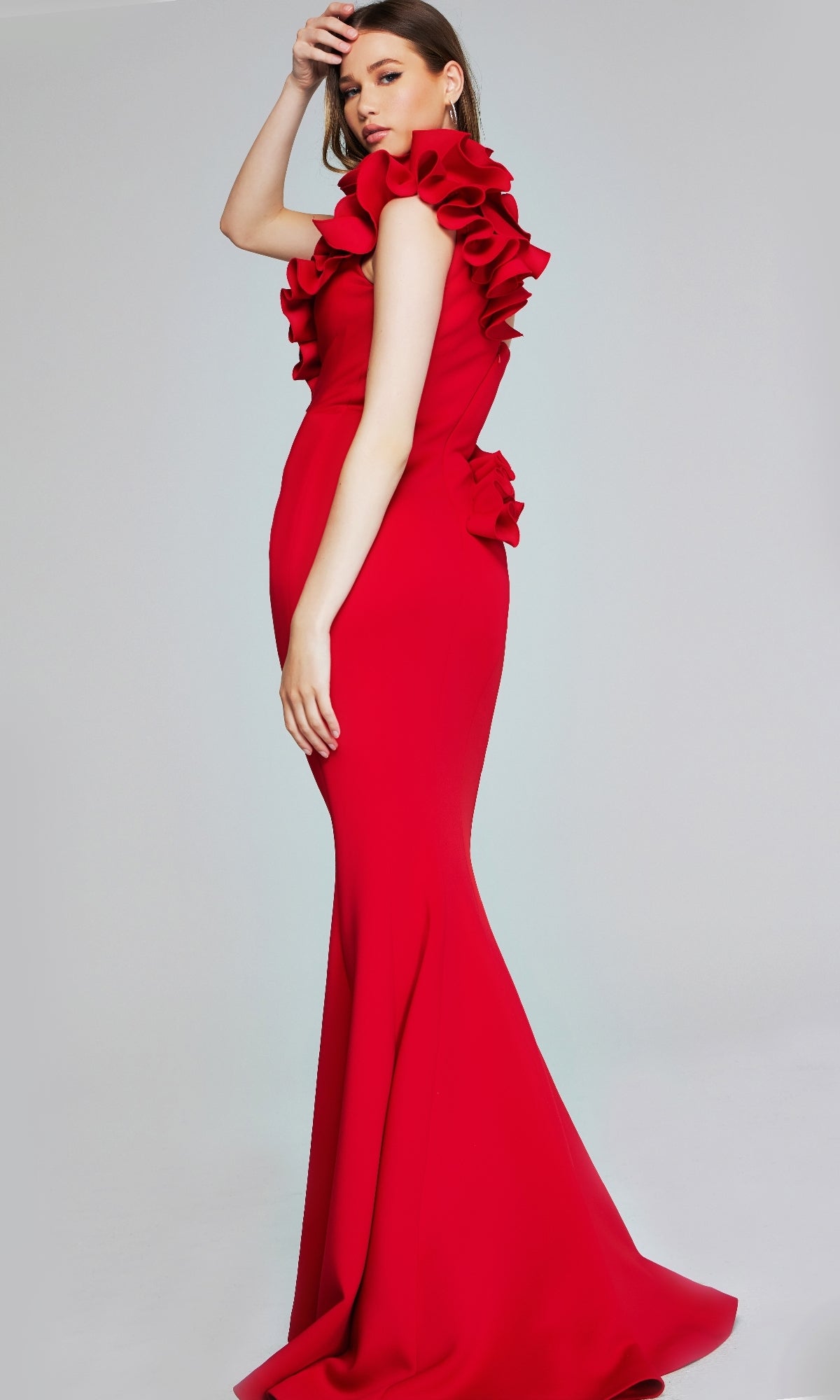 A woman poses against a plain light gray background wearing the Jovani 39751, a long formal fitted red mermaid gown with ruffled details on the shoulders and back.