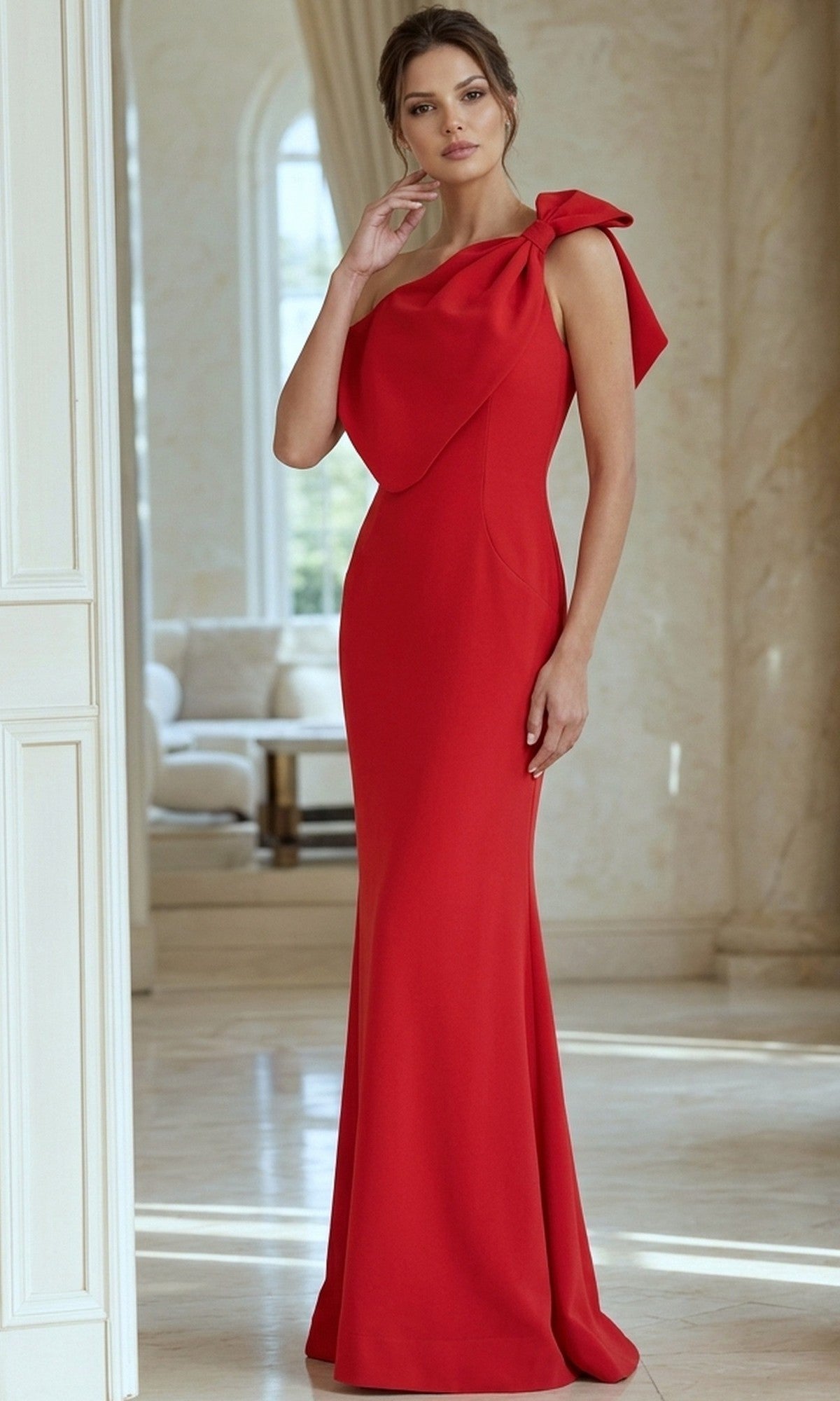 A woman stands indoors in a bright, elegant room wearing the Jovani 39971, a long formal dress featuring a fitted red evening gown with an asymmetric neckline and a bow detail on one shoulder.