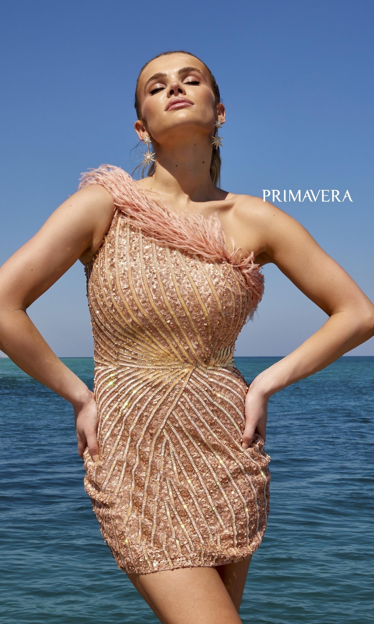 A woman stands by the ocean in the One-Shoulder Sequin Party Dress with Feathers 4002, featuring gold and pink hues, a textured sequin design, and feather-trimmed bodice.