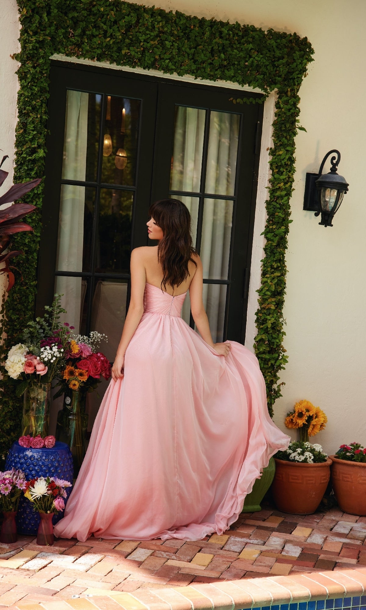 A woman wearing the Ava Presley 40041 A-Line Long Strapless Prom Dress stands outside glass doors, holding her gown amid colorful flower arrangements in pots and vases.