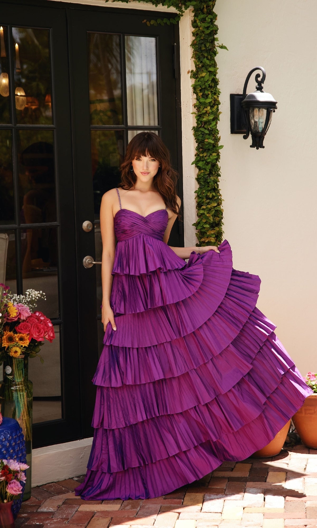 A person stands before glass doors, holding the edge of the Ava Presley 40046 Ruffled Prom Ball Gown with a sweetheart neckline. Nearby, flowers in vases are arranged on the ground.