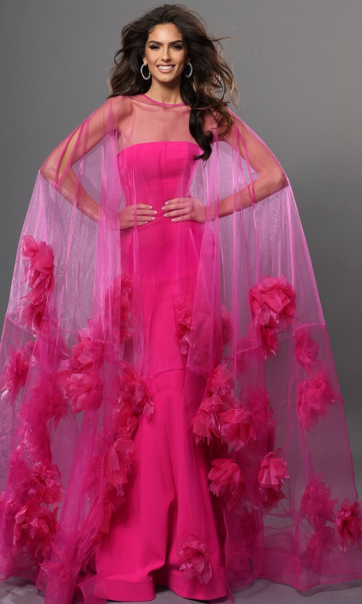A woman in a vibrant fuchsia Jovani 40092 long formal dress, featuring a sheer matching cape embellished with bold pink floral appliqués, stands against a plain gray background. This exquisite designer gown radiates elegance and sophistication.