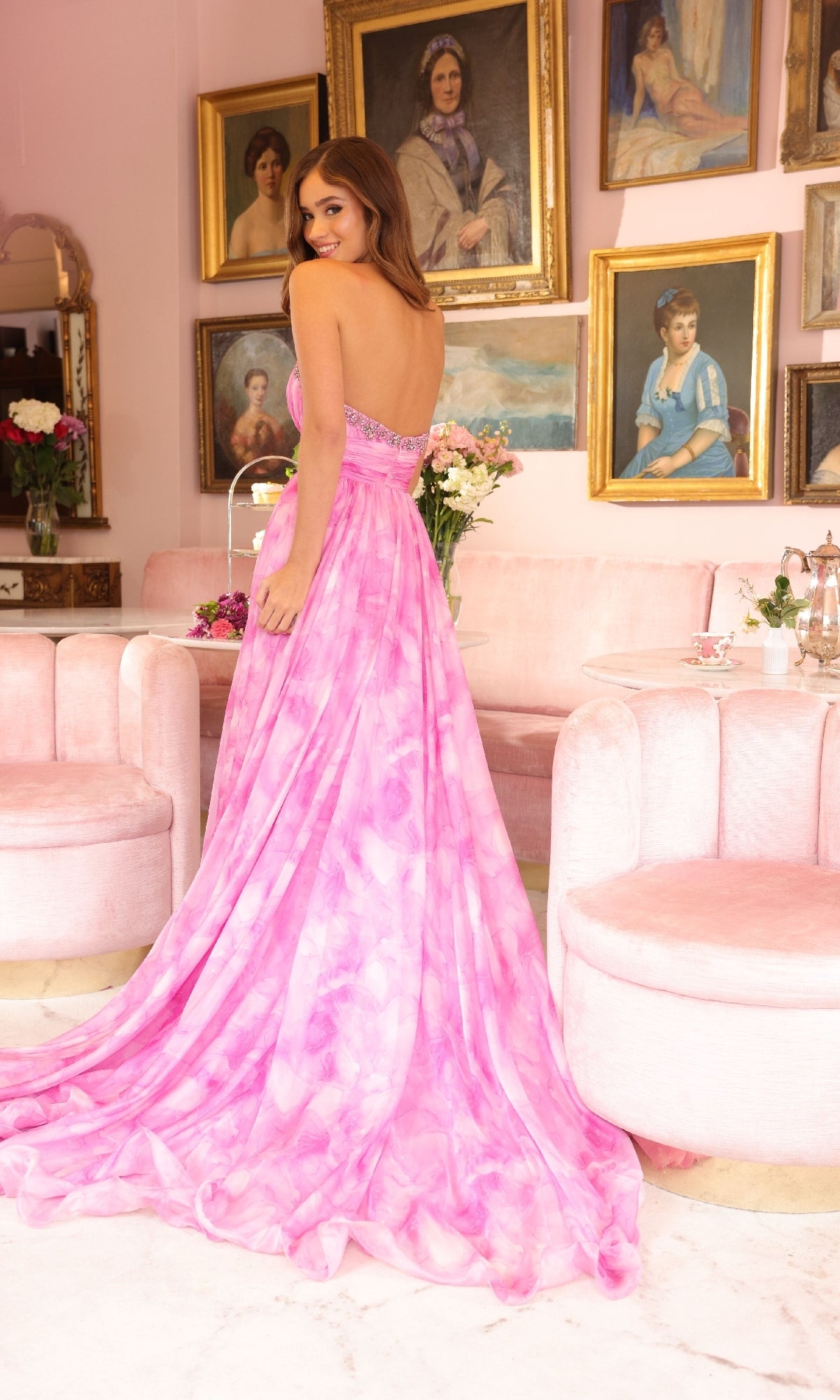 A woman wearing the Ava Presley 40116 Strapless Tie-Dye Prom Dress stands in a room with light pink furniture and portrait paintings on the wall.