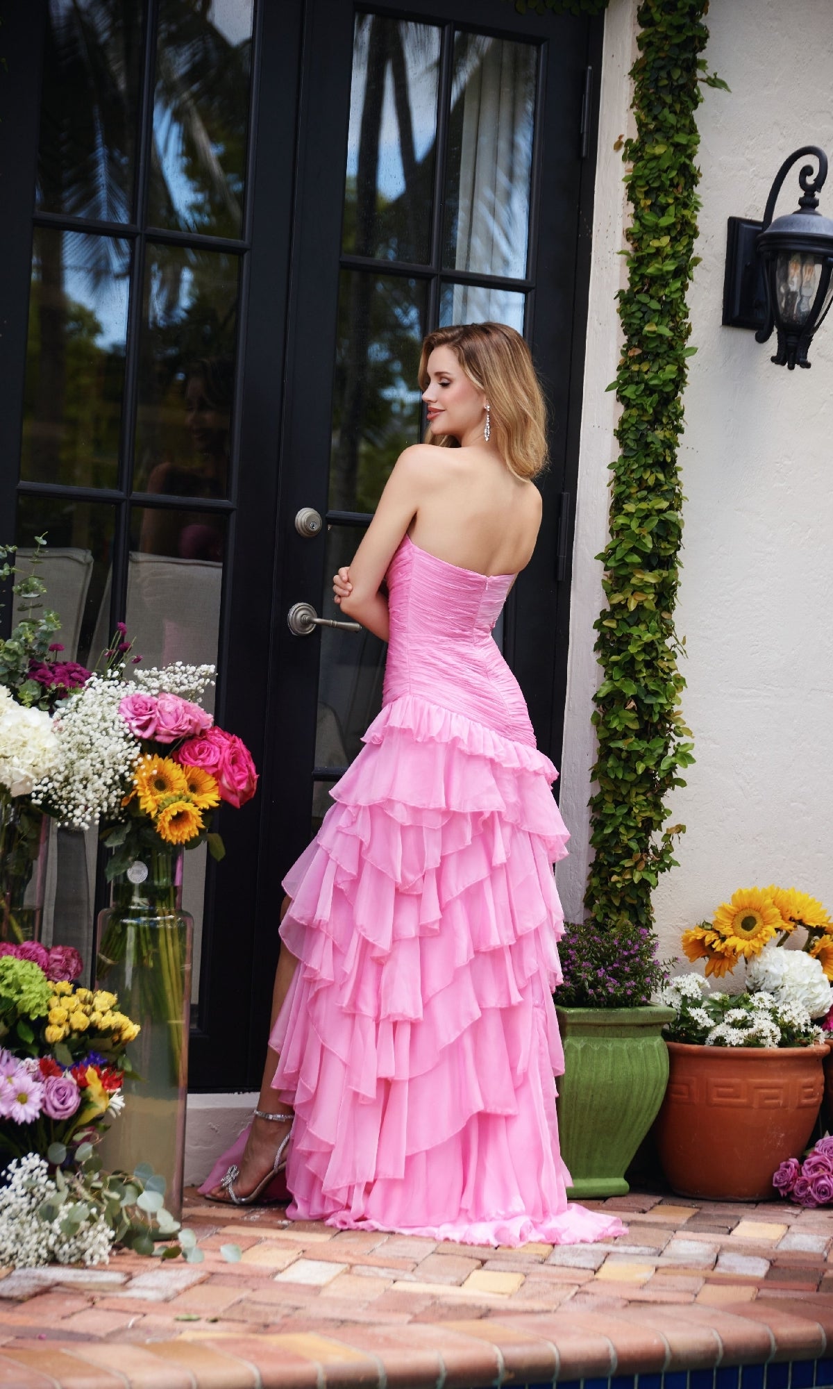 A woman wearing the Strapless Long Ruffle Prom Gown Ava Presley 40118 stands by a glass door on a tiled patio, surrounded by potted flowers and plants.