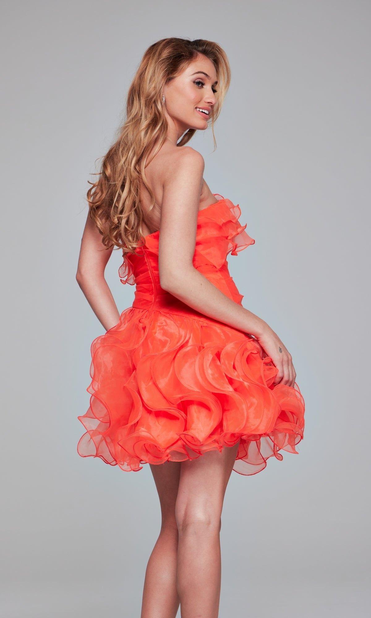 A woman stands sideways and smiles in the Jovani 40144 Strapless Short Ruffled Hoco Dress, featuring a bright orange color and A-line ruffled skirt, against a plain background.