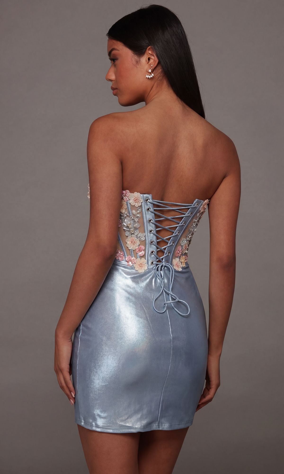 A woman faces away, modeling the Alyce 40145 short homecoming dress—a strapless, metallic blue design featuring a lace-up back and delicate floral embroidery on the corset-style bodice—while standing against a plain gray background.
