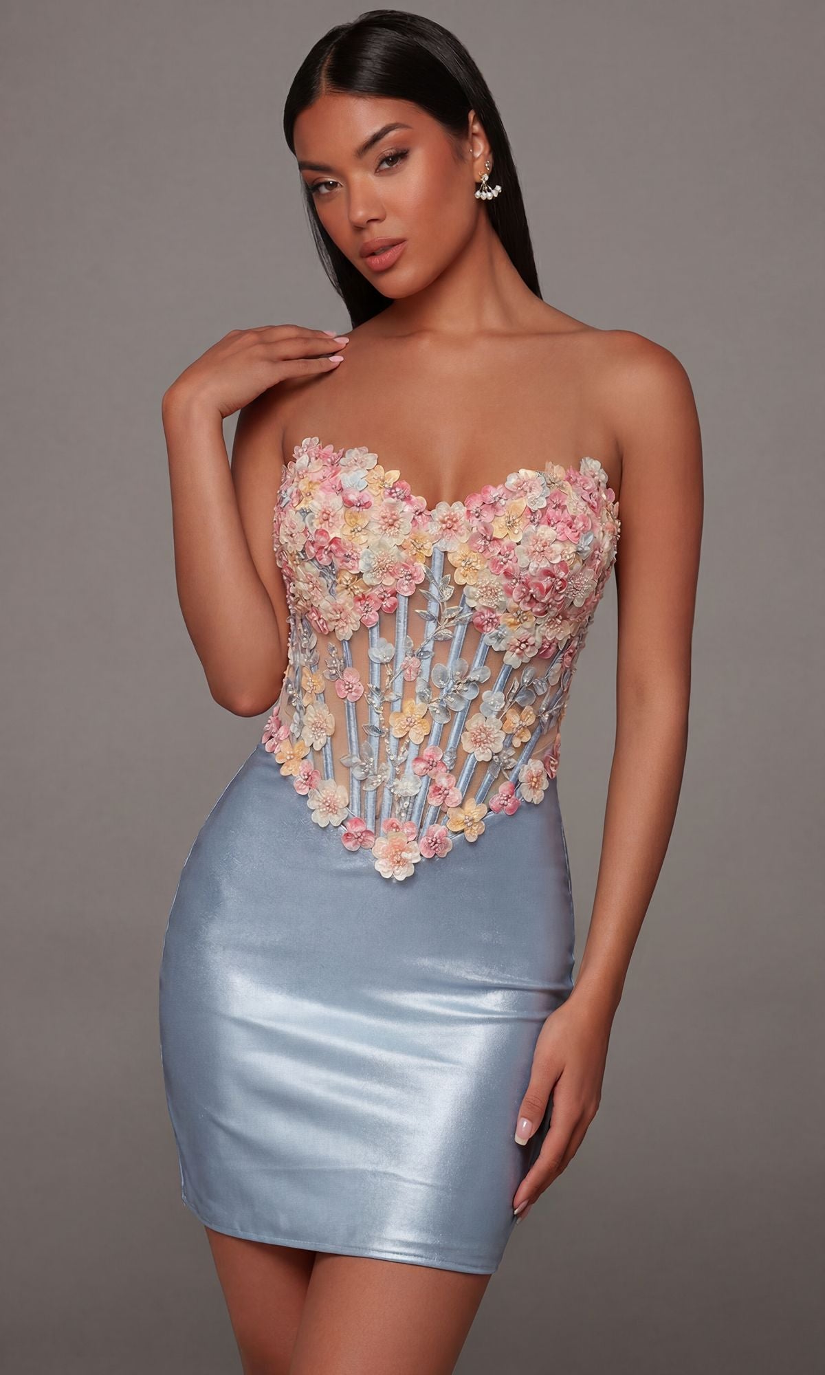 A woman wearing the Alyce 40145 short homecoming dress in French Blue, which features a strapless design with pastel floral appliqué details on the bodice reminiscent of a floral embroidered corset, stands against a plain gray background.