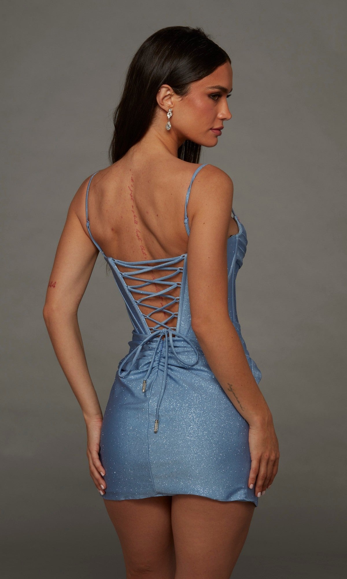 A woman is seen from behind wearing the Alyce 40146 short homecoming dress, featuring a sparkly light blue fabric, lace-up back, and corset bodice, standing against a plain gray background.