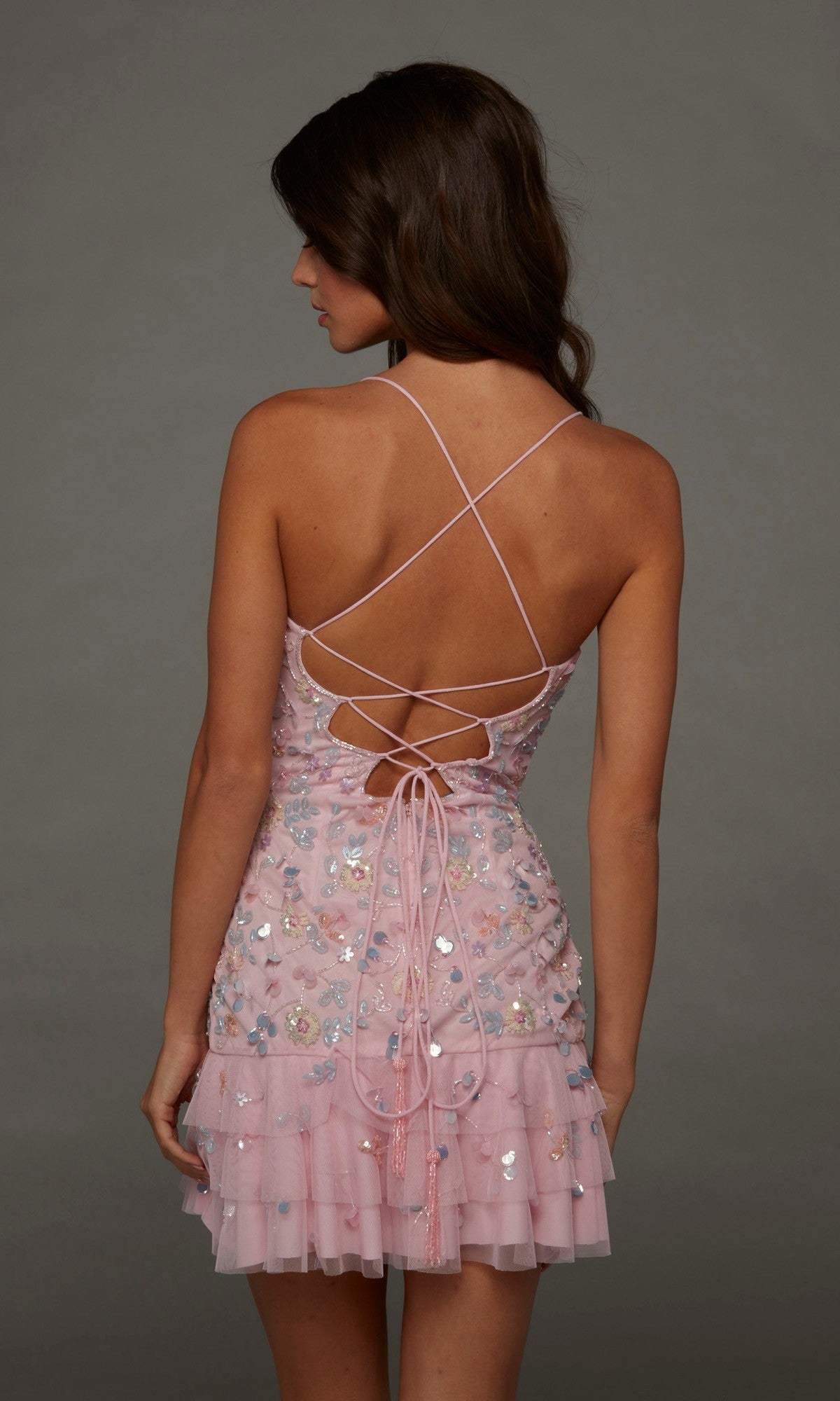 Woman wearing the Alyce 40148 Short Homecoming Dress, featuring pink strappy sequins, beaded embellishments, and a ruffled hem, shown from the back against a plain gray background.