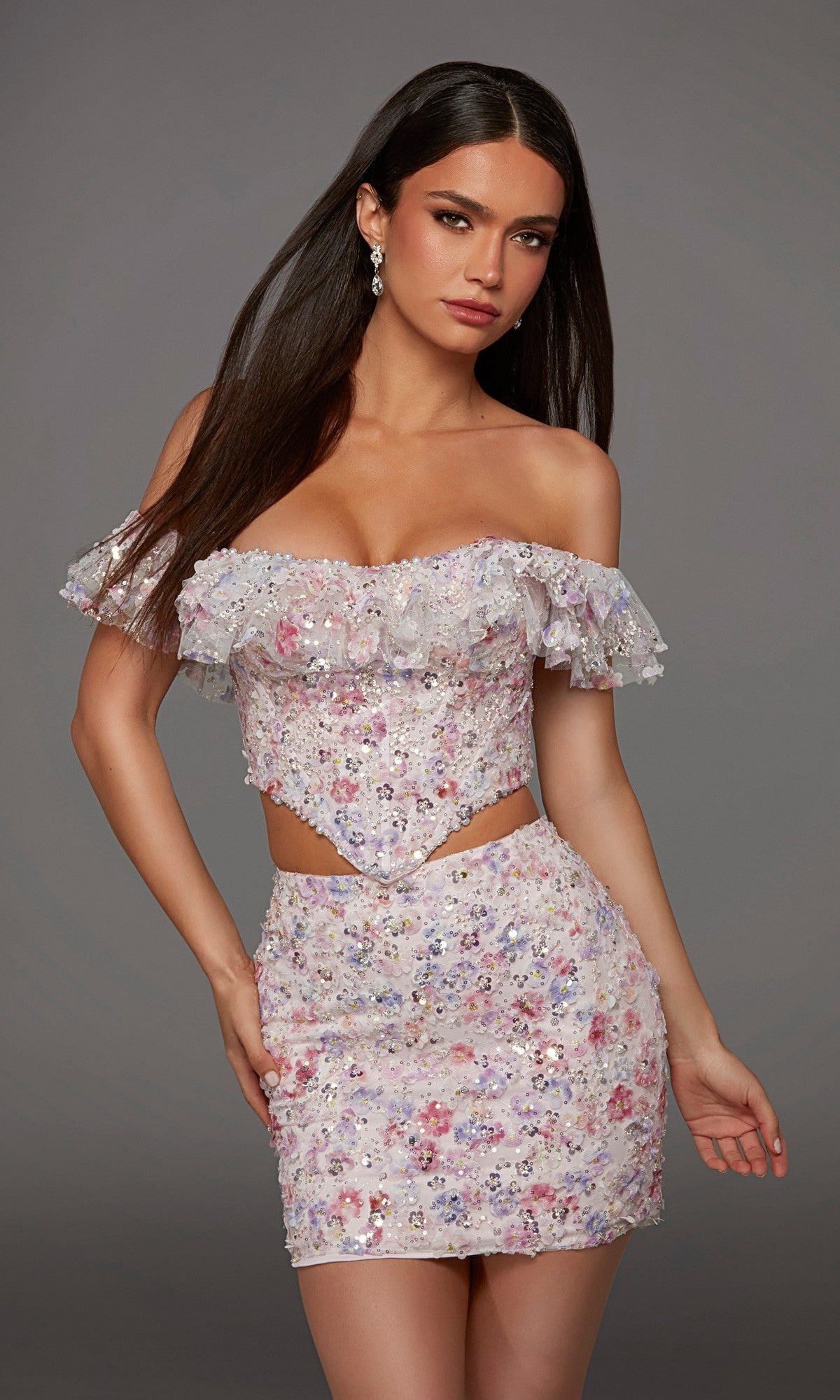 A woman stands against a plain gray background wearing the Alyce 40167 short homecoming dress—a pastel, off-shoulder crop top embellished with 3D floral embroidered appliques and sequins, paired with a coordinating mini skirt.