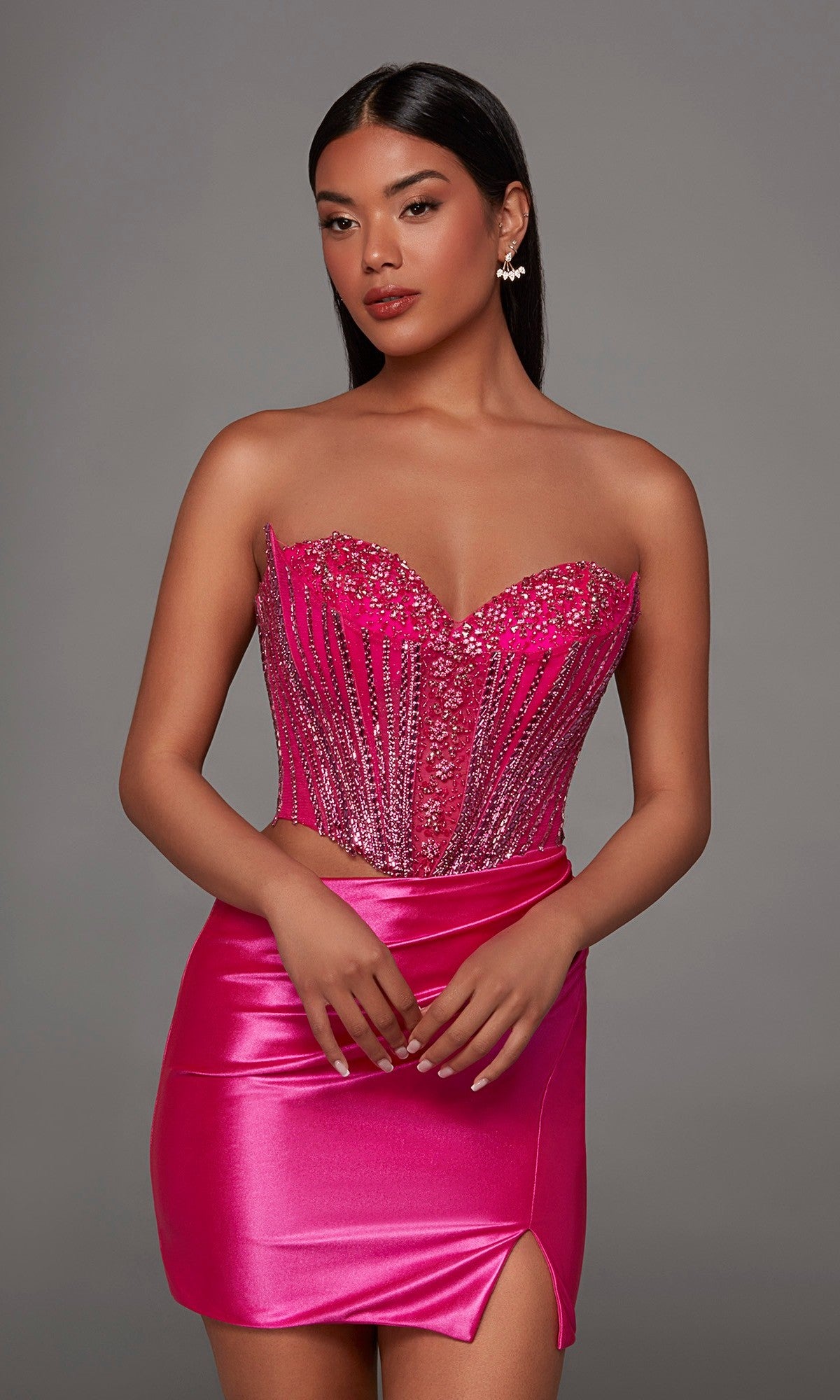 A woman wearing the Alyce 40194 short homecoming dress in Barbie Pink, which showcases a strapless design with an embellished corset bodice and sparkling beading, paired with a shiny satin short skirt featuring a thigh-high slit, stands against a plain gray background.