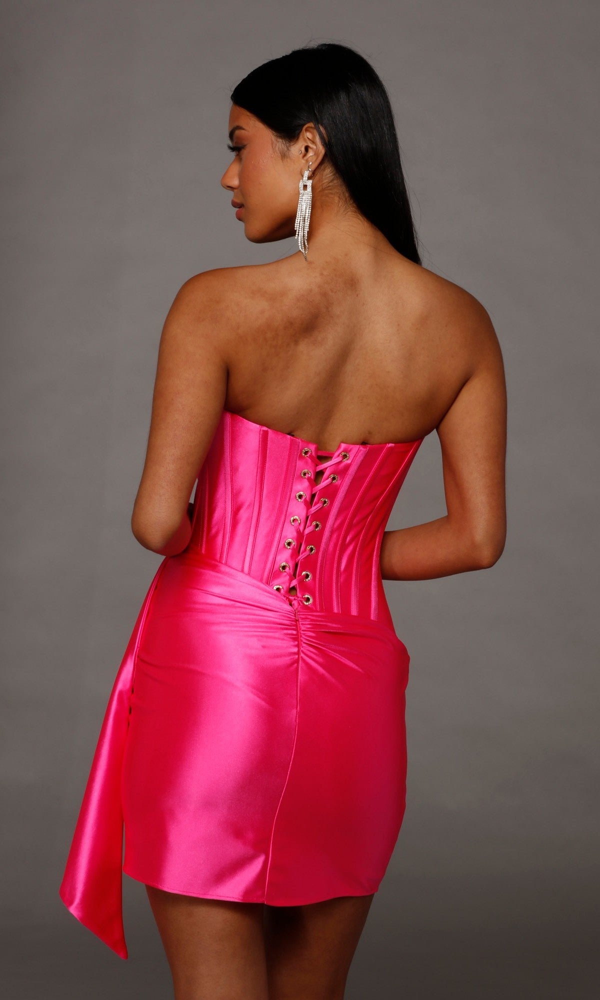 A woman wearing the Alyce 40207 short homecoming dress in bright pink stretch-satin, showcasing its strapless design and corset bodice with lace-up back, stands against a plain gray background, facing away from the camera.