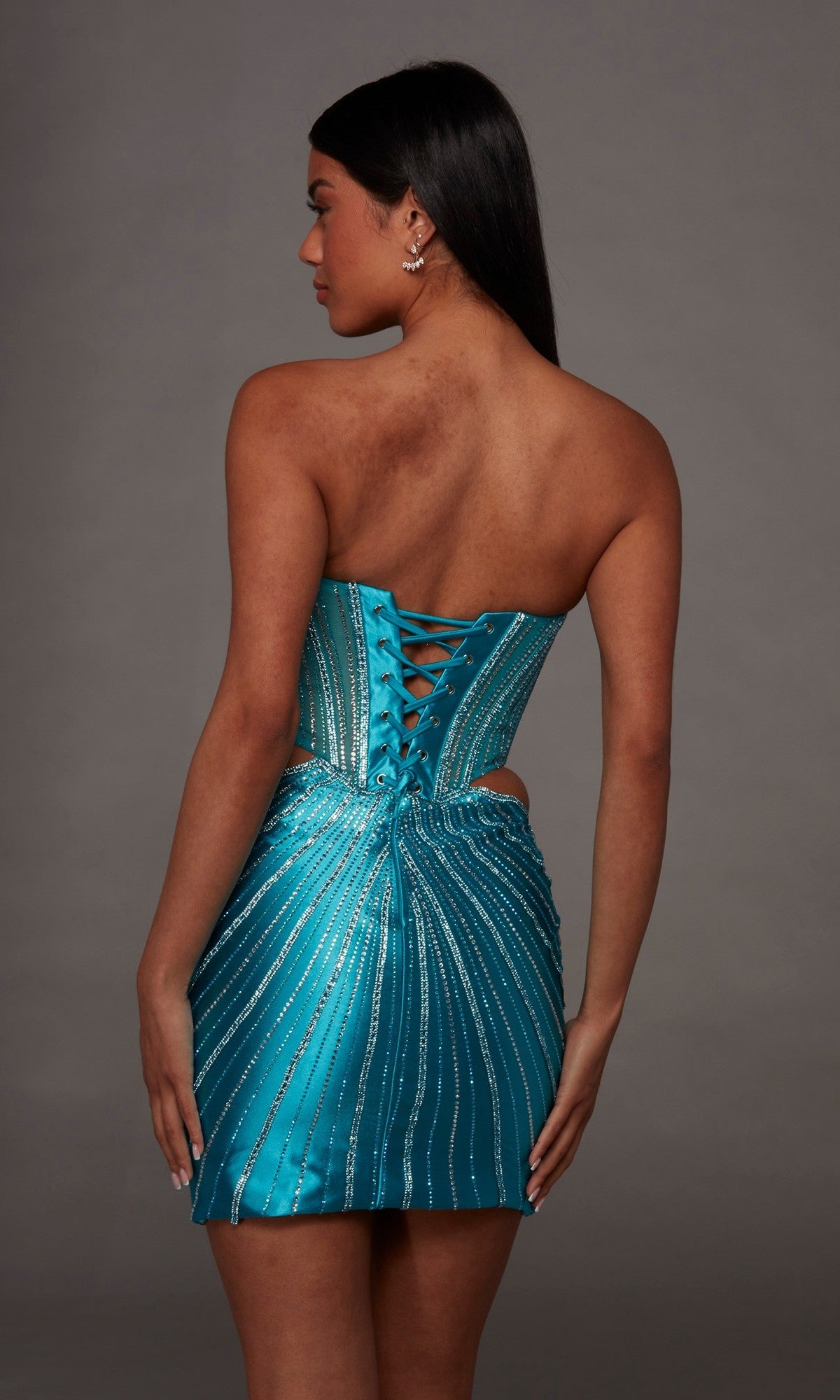 Model wearing the Alyce 40211 Short Homecoming Dress—a strapless, teal mini dress with silver sparkly stripes, a lace-up back, and a flattering corset bodice—posed with her back to the camera against a plain gray background.