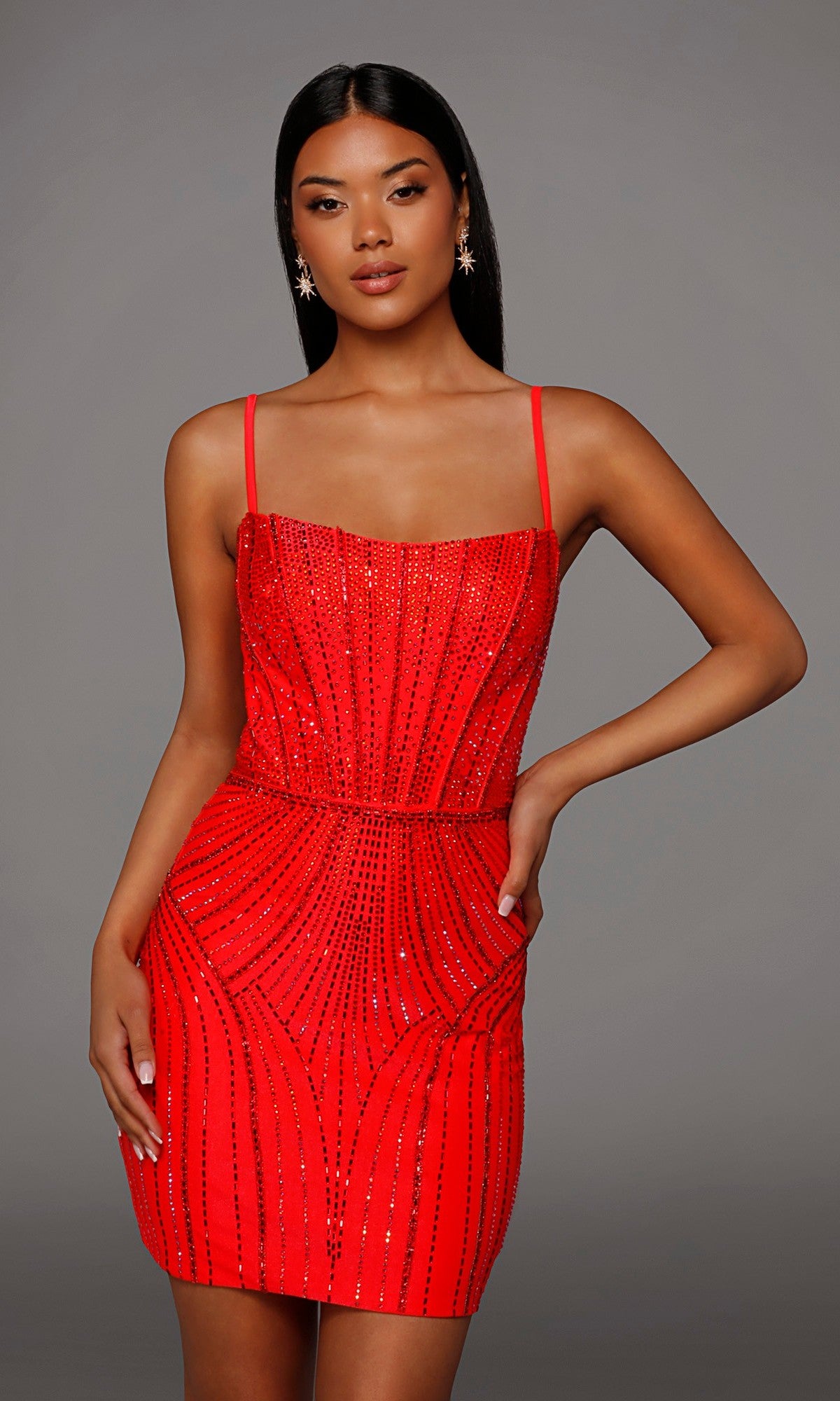 A woman wearing the Alyce 40220 short homecoming dress in watermelon, featuring a fitted silhouette with delicate beading and thin straps, stands against a plain gray background with one hand on her hip.