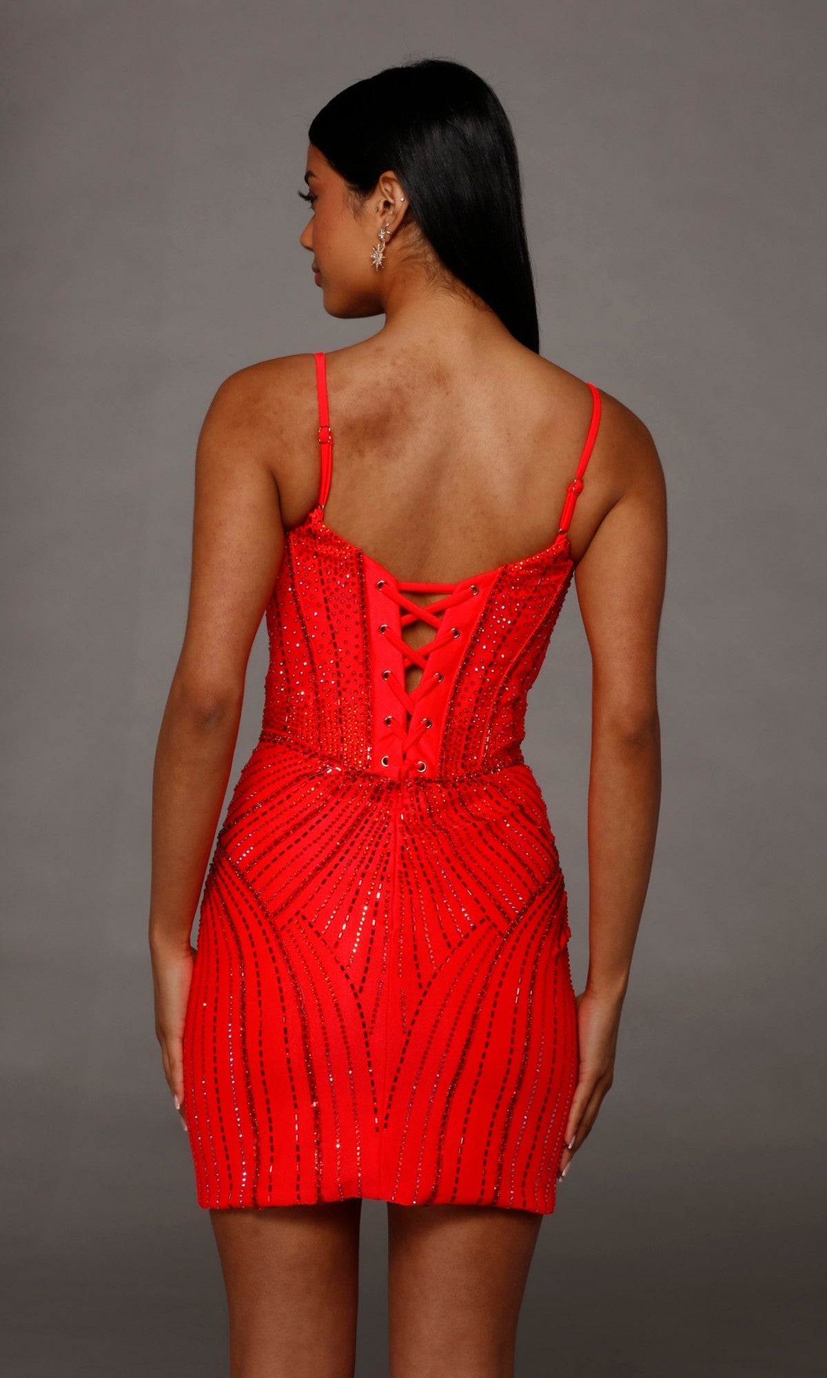 A woman facing away showcases the Alyce 40220 Short Homecoming Dress—a red, beaded, strappy mini fitted dress with a lace-up back—against a plain gray background.