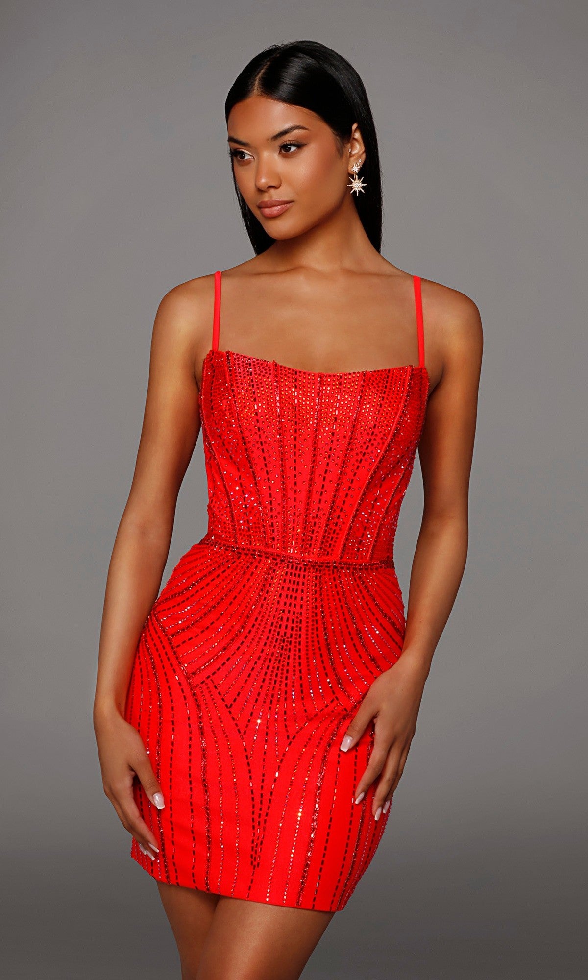 A woman wearing the Short Homecoming Dress: Alyce 40220—a fitted red dress with thin straps and sparkling rhinestone embellishments—stands against a plain gray background.
