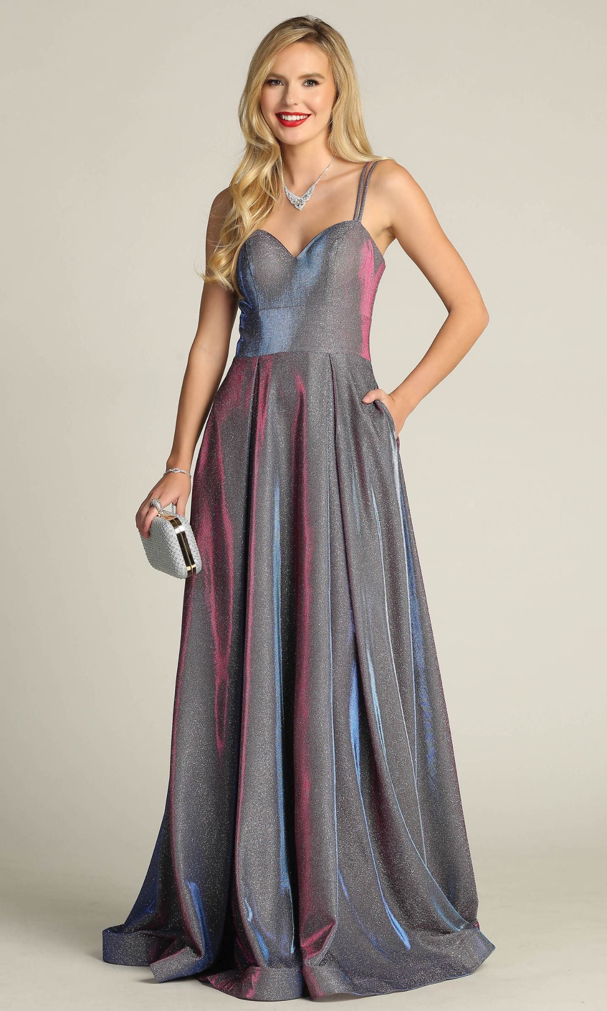 A woman in the Lace-Up Back Long Metallic A-Line Prom Dress 4022, featuring a shimmering silver-gray finish and thin straps, holds a small clutch while standing against a plain background.