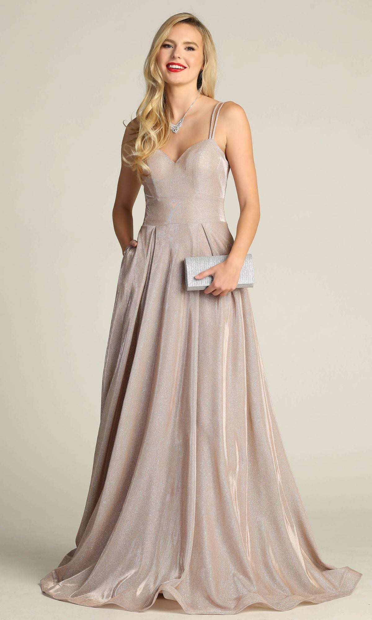 A woman in the Lace-Up Back Long Metallic A-Line Prom Dress 4022, a shimmering floor-length champagne gown with thin straps and a sweetheart neckline, holds a textured clutch and smiles at the camera.