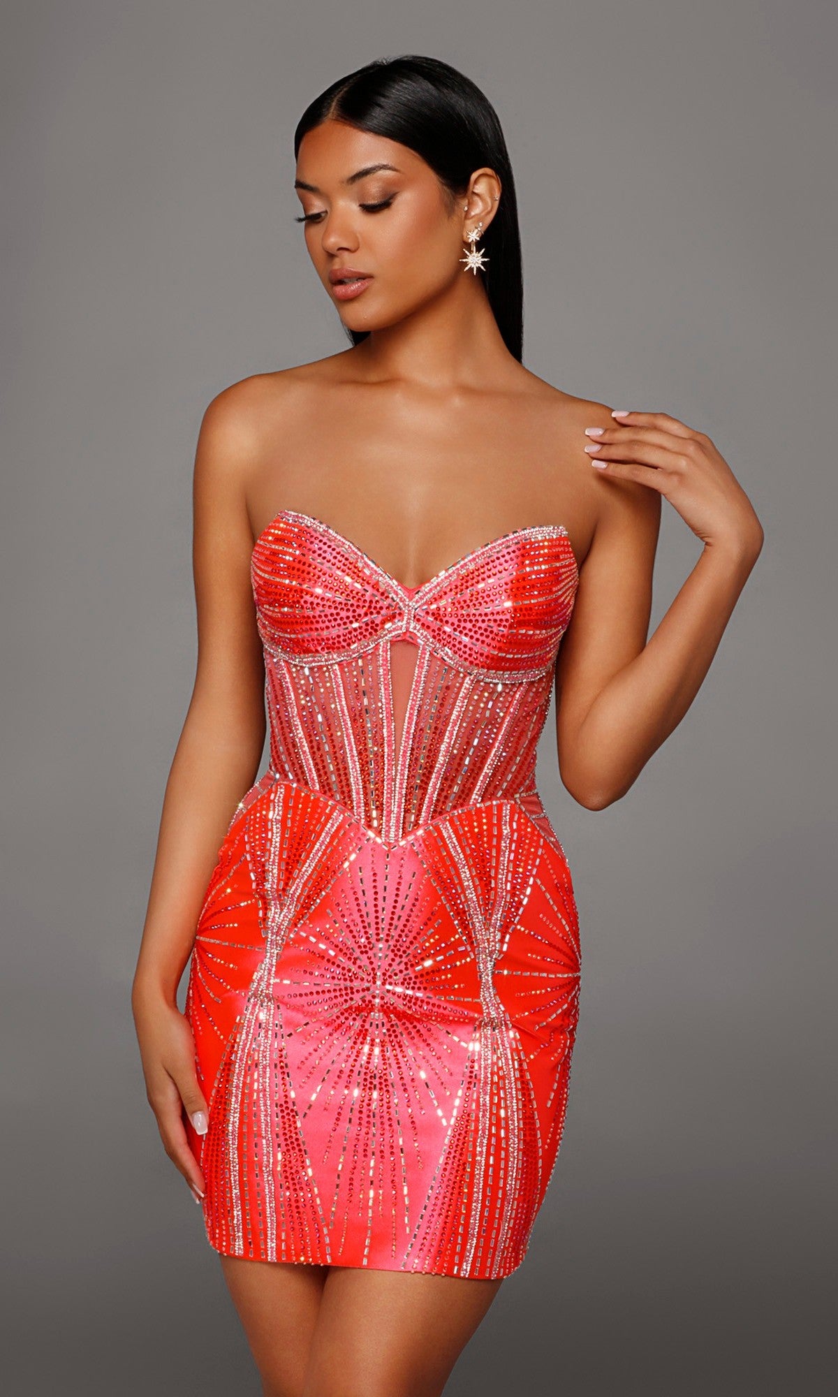 A woman poses against a gray background in the Alyce 40232 short homecoming dress, a fitted strapless mini in hot coral featuring geometric patterns and sparkling rhinestone embellishments.