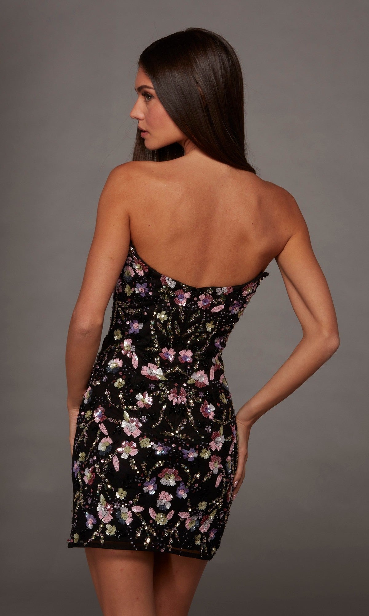 A woman poses with her back to the camera, modeling the Alyce 40239 Short Homecoming Dress—a strapless, fitted black dress featuring a vibrant floral pattern and delicate sequin accents—set against a plain gray background.