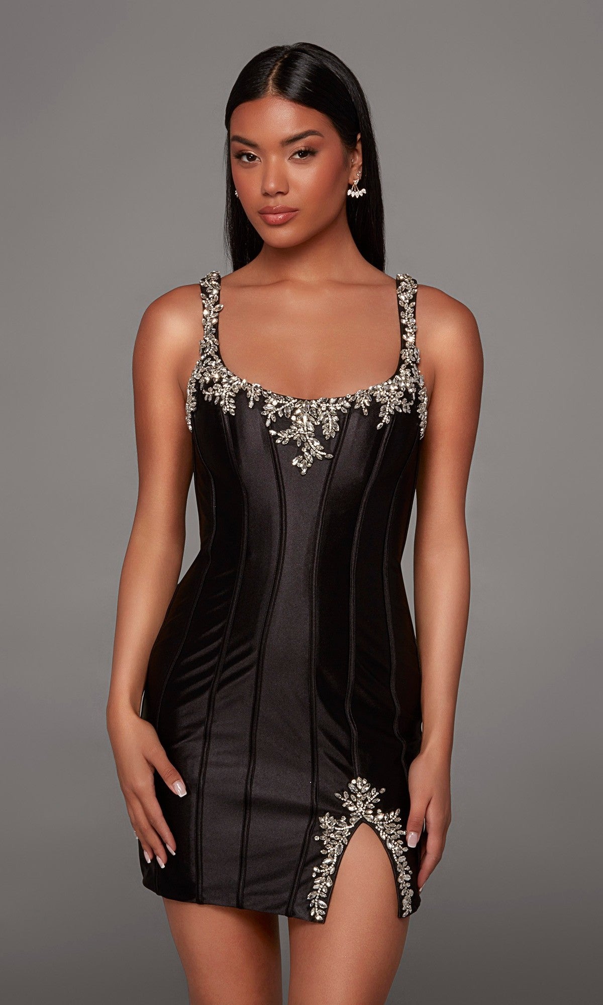 A woman models the Short Homecoming Dress: Alyce 40243 in Black/Silver, featuring a fitted satin silhouette with metallic floral embellishments on the straps, neckline, and skirt slit, standing against a plain gray background.