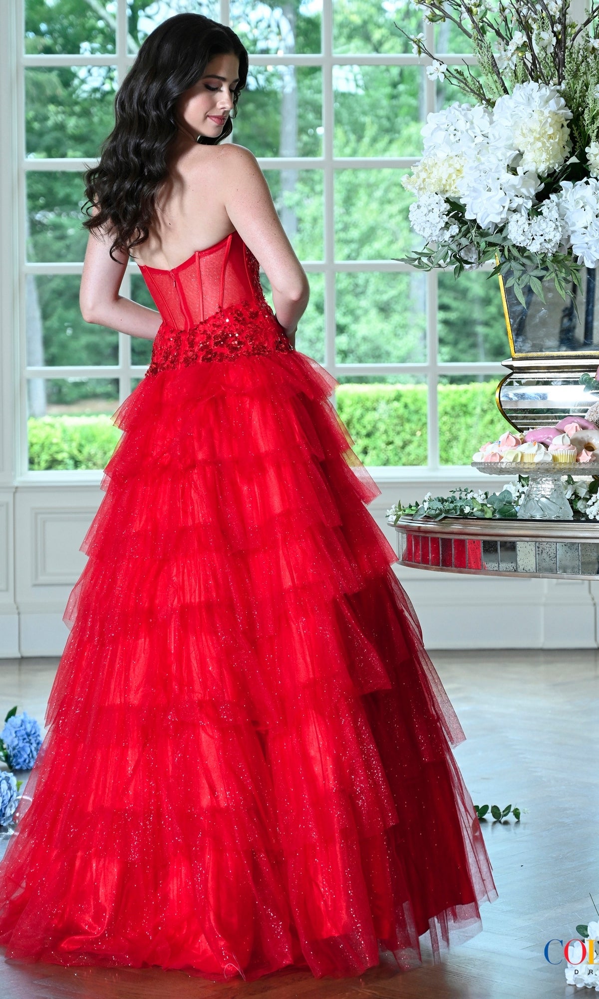 A woman wears a Colors Dress 4024 strapless red glitter tulle ball gown with a corset bodice and a tiered skirt while standing near a window.