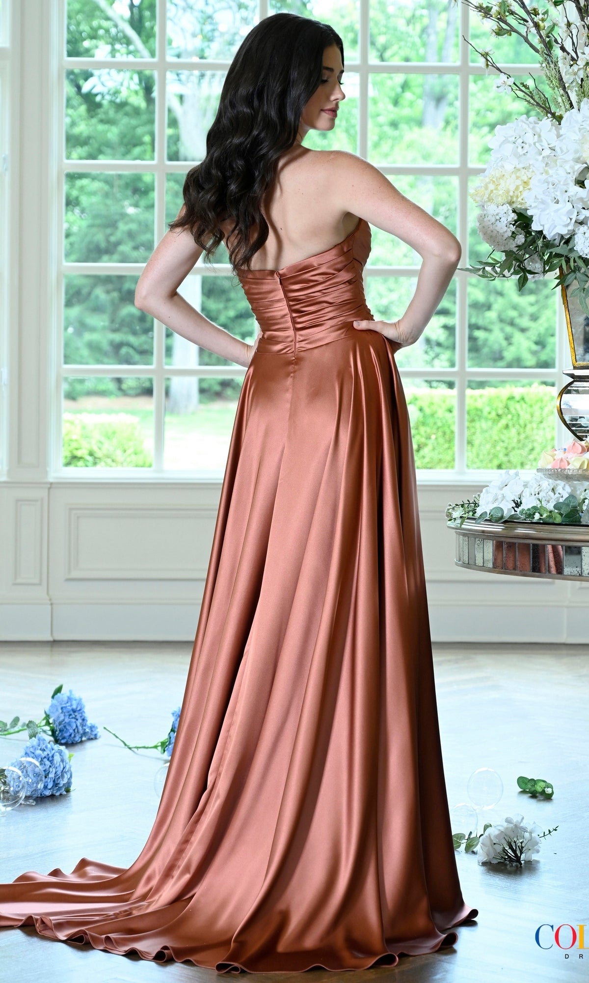 A woman wears a copper-colored satin Long Prom Dress: Colors Dress 4026, featuring a strapless, old Hollywood-style silhouette as she stands with her back to the camera in front of a window.