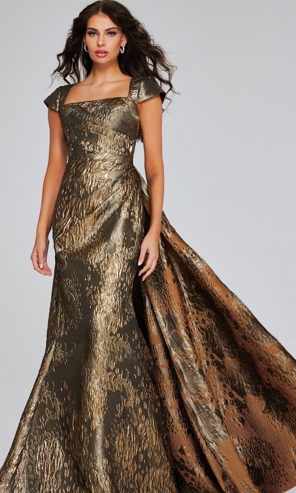 A woman wears the Long Formal Dress: Jovani 40295, an elegant metallic gold and bronze textured gown with a dramatic train, standing against a plain light background.