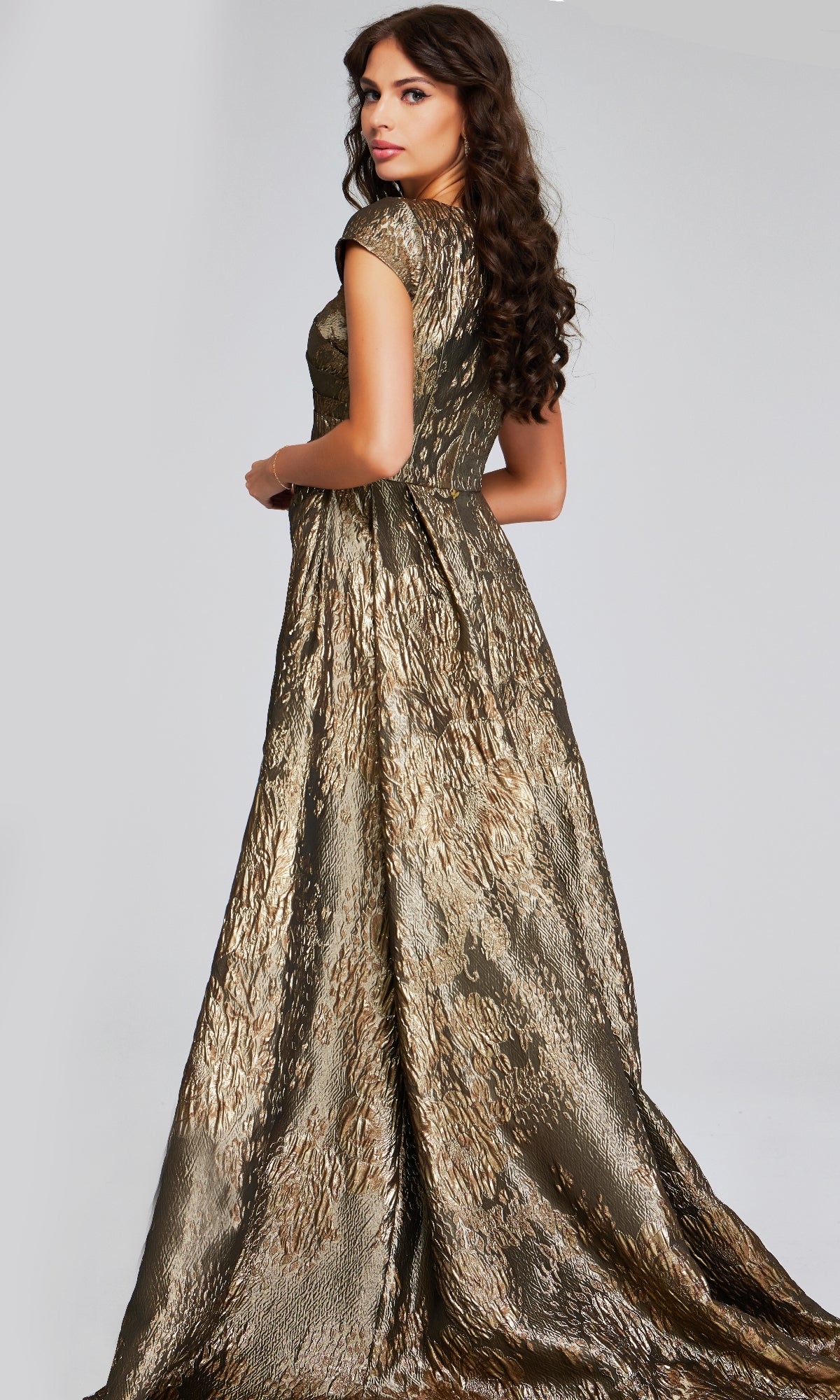 A woman with long, wavy brown hair wears the gold, textured Jovani 40295 long formal dress with short sleeves and a floor-length silhouette, standing against a plain light gray background.