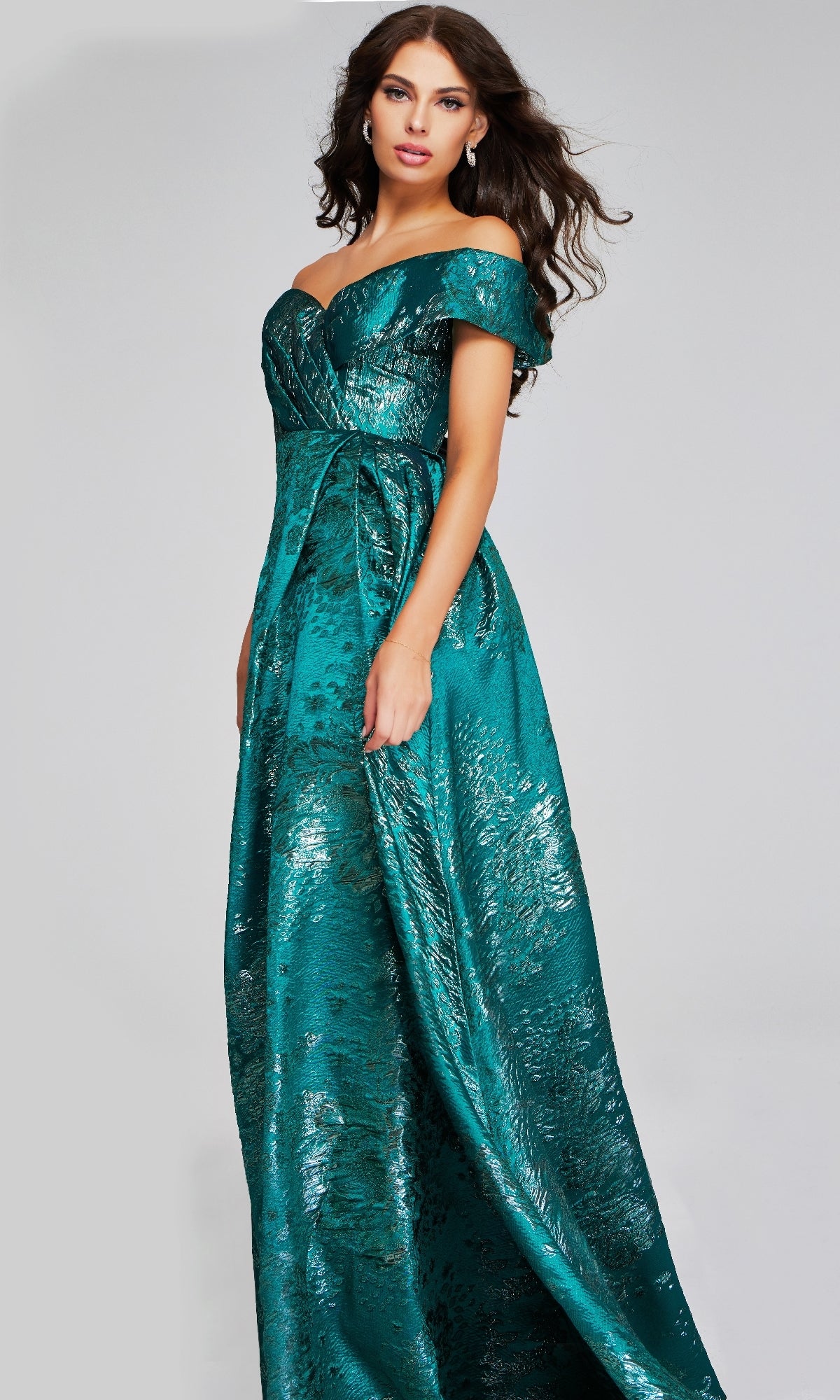 Woman wearing the Jovani 40296 long formal dress—an off-the-shoulder, floor-length gown in metallic teal textured fabric—stands against a plain light background.