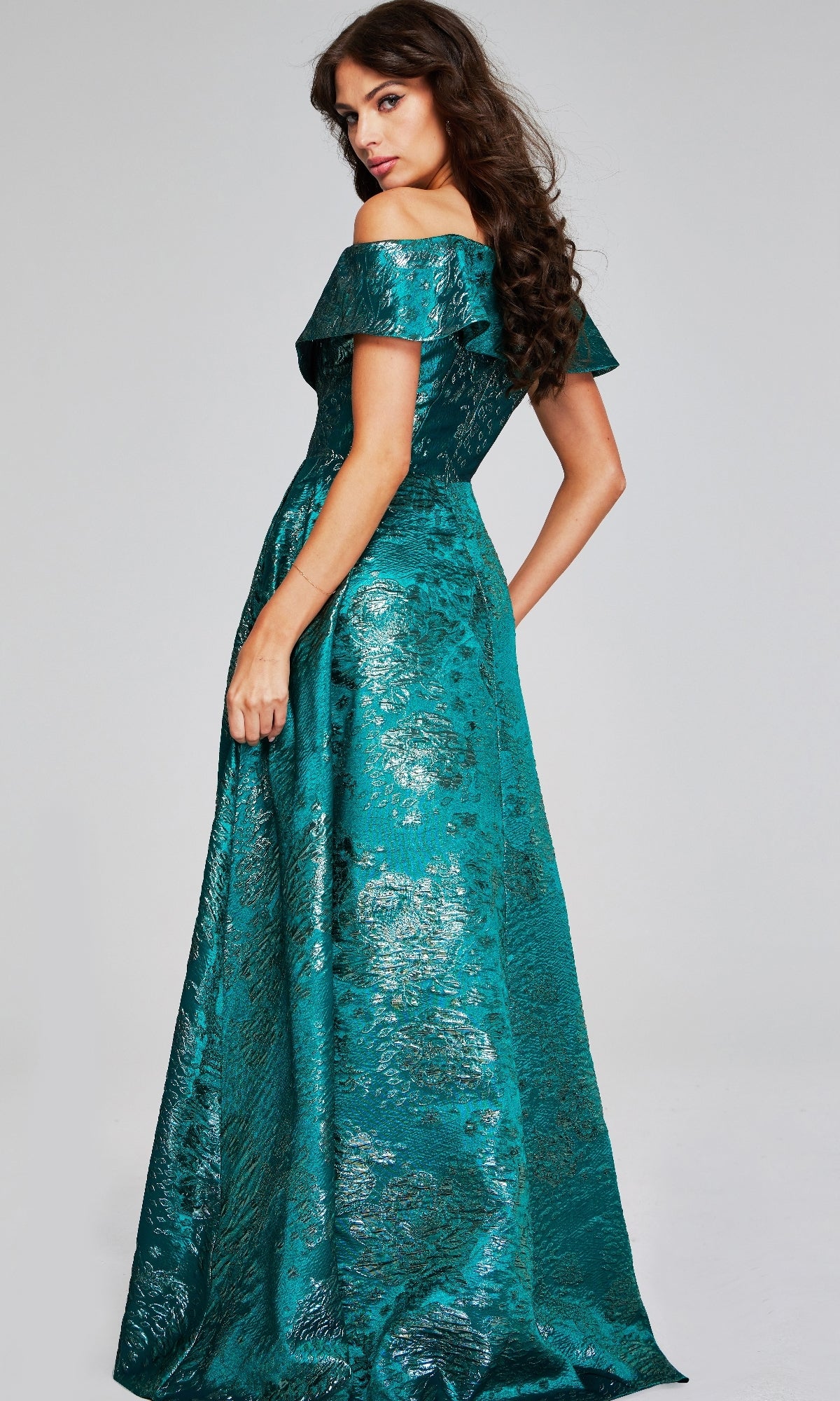 A woman with long brown hair poses in the Jovani 40296 long formal dress, featuring off-the-shoulder styling and a shimmery teal metallic textured fabric, set against a plain background.