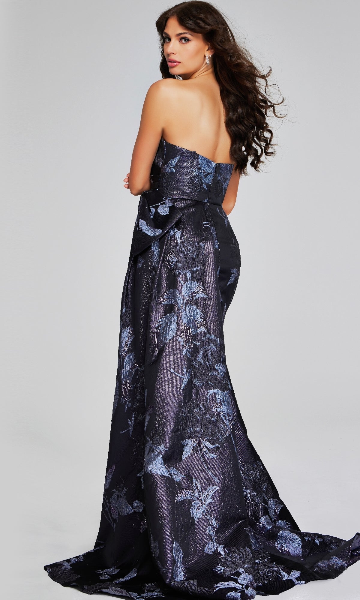 A woman showcases the Jovani 40308 long formal dress—an elegant, strapless, floor-length gown in dark blue floral jacquard with a textured pattern—as she stands turned away, glancing over her shoulder.