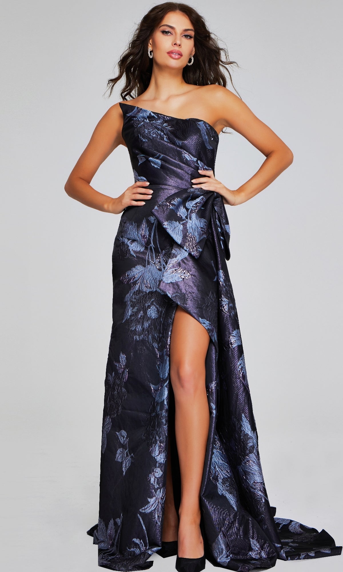 A woman poses in the Jovani 40308 long formal dress, featuring a dark floral jacquard pattern, thigh-high slit, and black high heels against a plain background.